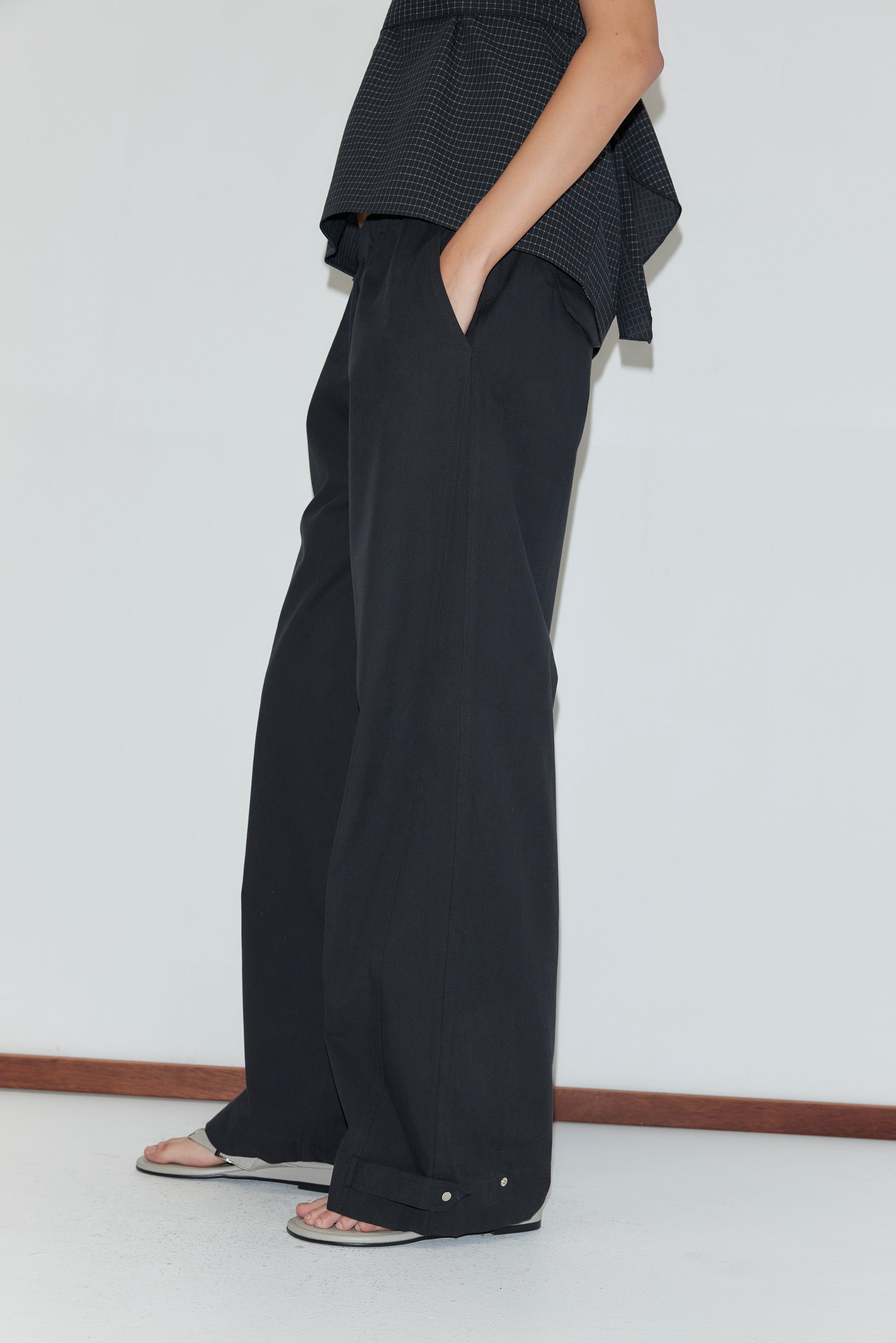 A person stands against a white wall wearing loose-fitting Snap Tab Pant - Black, a dark patterned top, and flip-flops. One hand is in their pocket, and their head is out of frame.