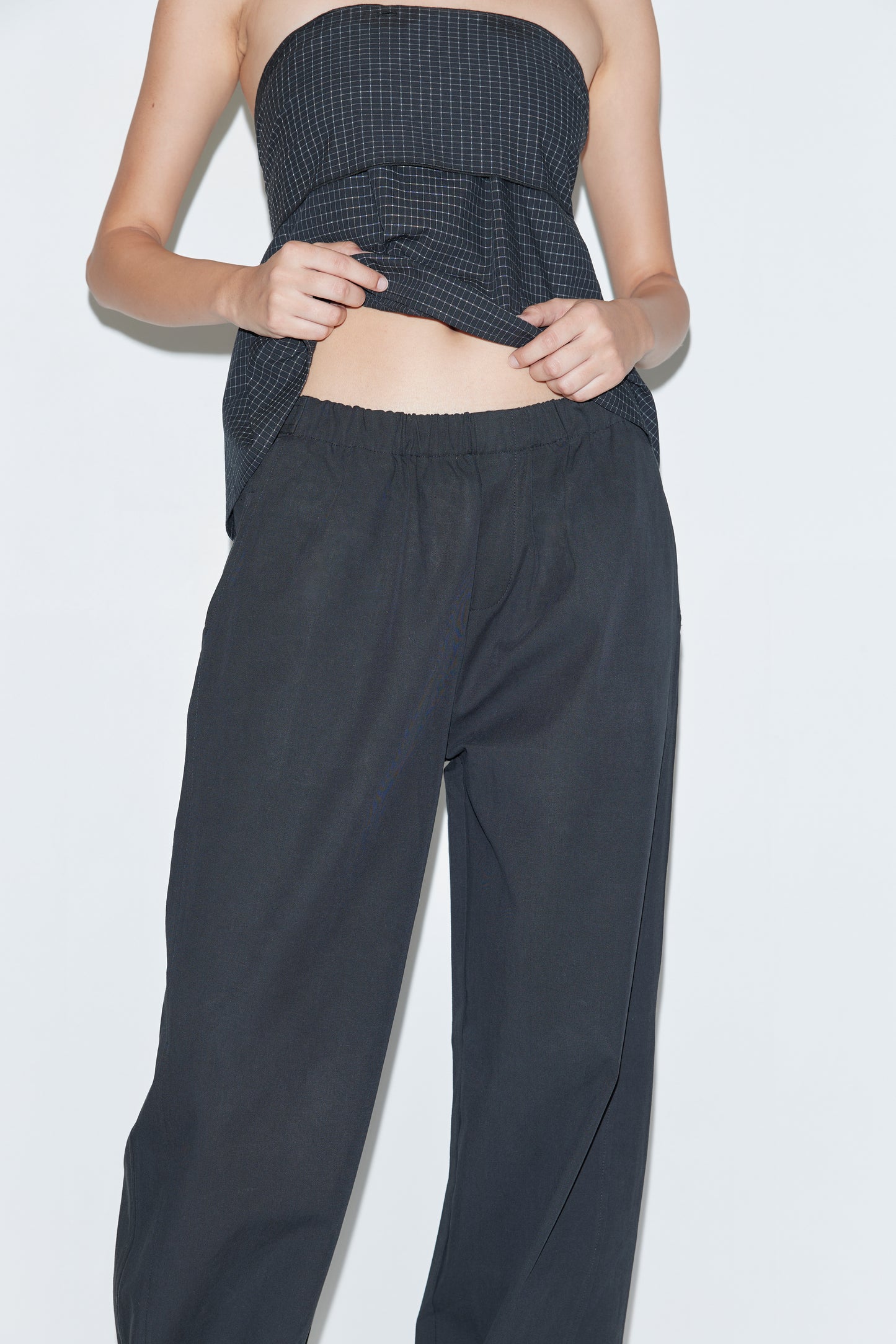 Wearing the Snap Tab Pant - Black, a person stands against a plain light background in relaxed wide-leg pants and a strapless textured dark top, lifting part of the top to reveal their midriff.