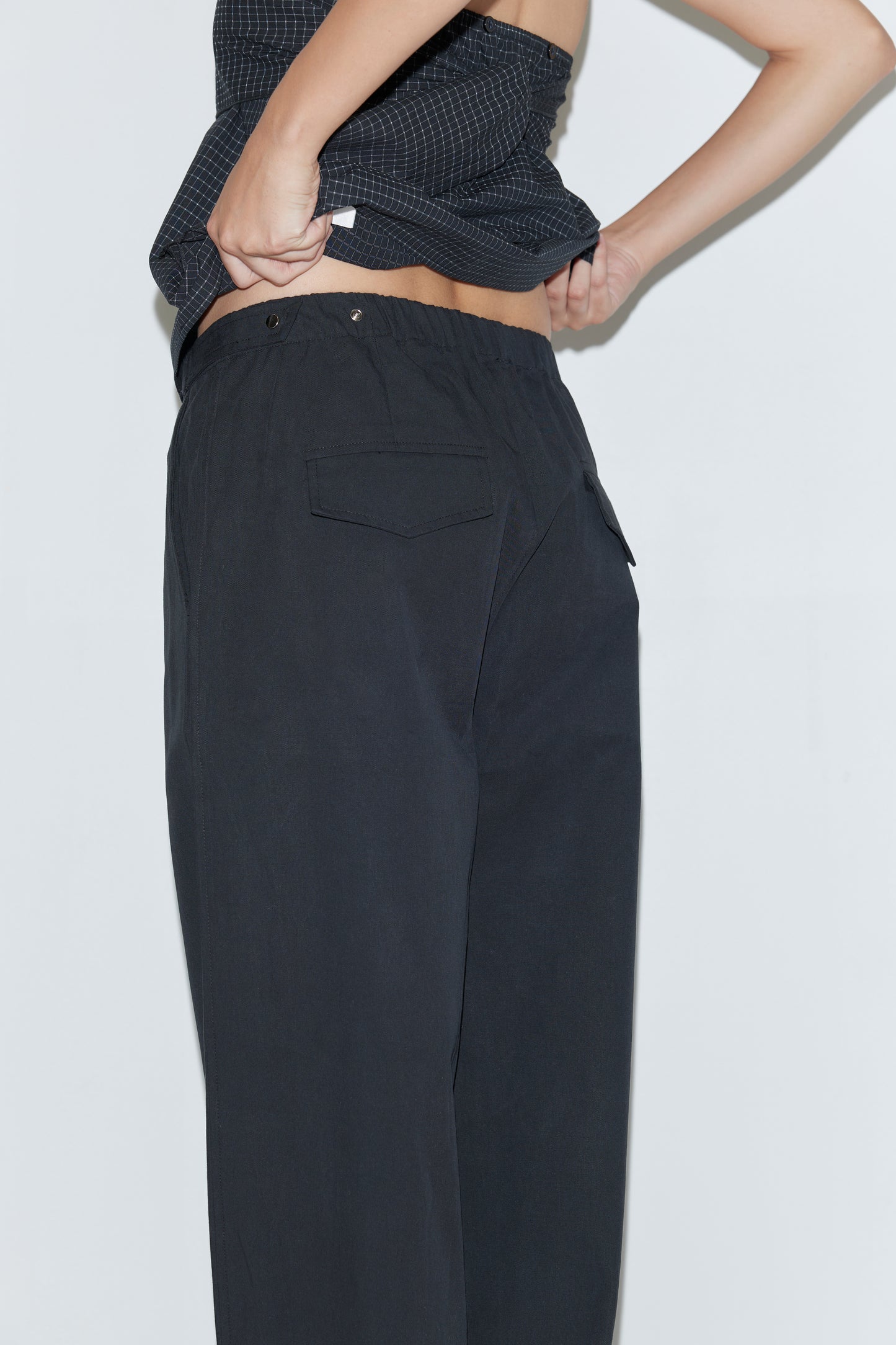 Wearing the Snap Tab Pant - Black, a person lifts their dark top to reveal part of their lower back, standing against a plain light background. The focus is on their midsection and the stylish Deiji Studios pants.