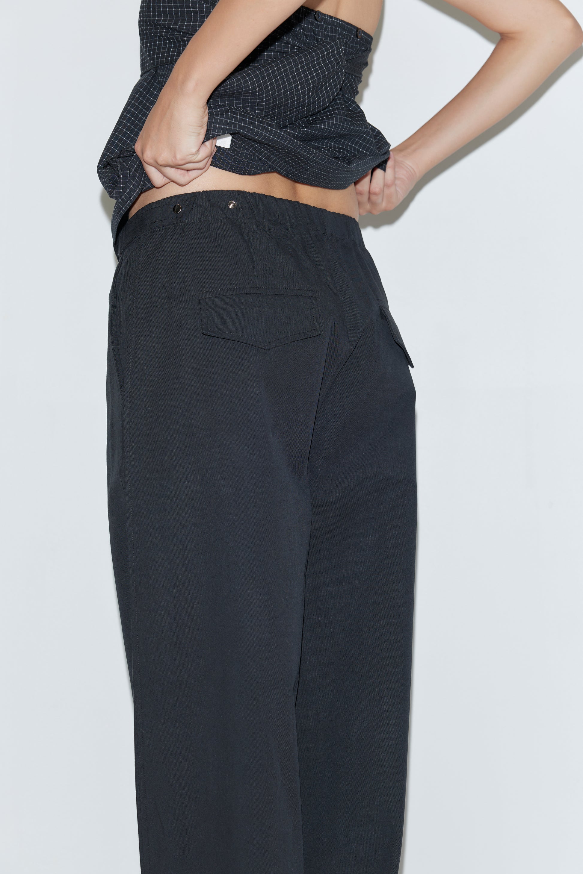 Wearing the Snap Tab Pant - Black, a person lifts their dark top to reveal part of their lower back, standing against a plain light background. The focus is on their midsection and the stylish Deiji Studios pants.