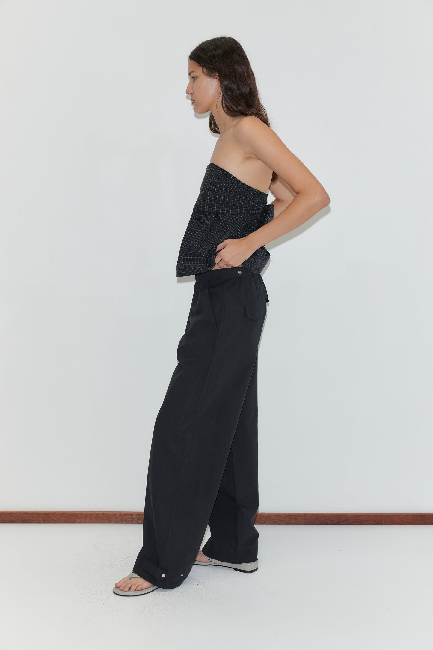 A person stands sideways against a white wall, wearing a strapless textured top, Snap Tab Pant - Black, and black sandals. Their long hair is down, with one hand casually tucked in their pocket.