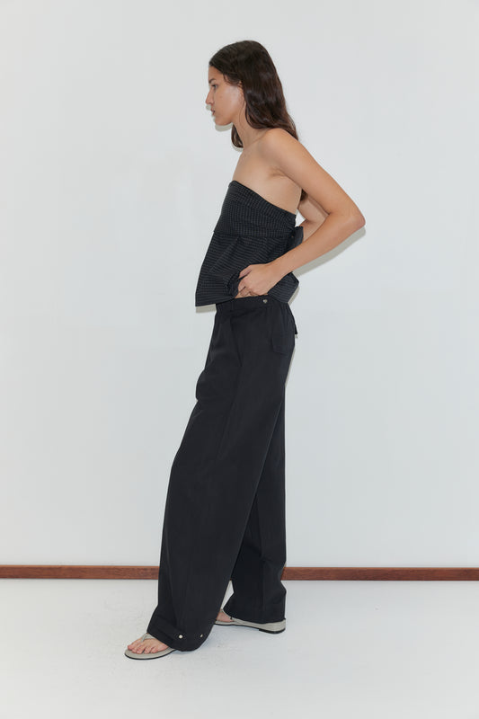 A person stands sideways against a white wall, wearing a strapless textured top, Snap Tab Pant - Black, and black sandals. Their long hair is down, with one hand casually tucked in their pocket.