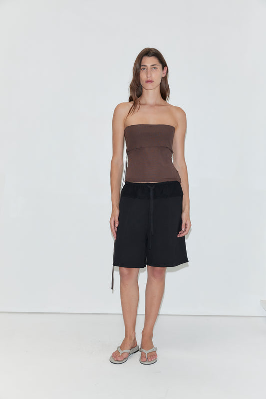 A woman with long brown hair and a neutral expression stands against a white wall, wearing a brown strapless top, Deiji Studios’ Contrast Mid Short in black, and flip-flops.