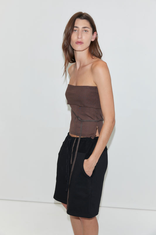 A woman with long, wavy brown hair poses against a white background, wearing a chocolate-brown ribbed strapless top with a tie-front detail and the Contrast Mid Short in Black by Deiji Studios, featuring relaxed jersey fabric and pockets. She has her hand tucked into her pocket, highlighting the effortless style of the shorts.
