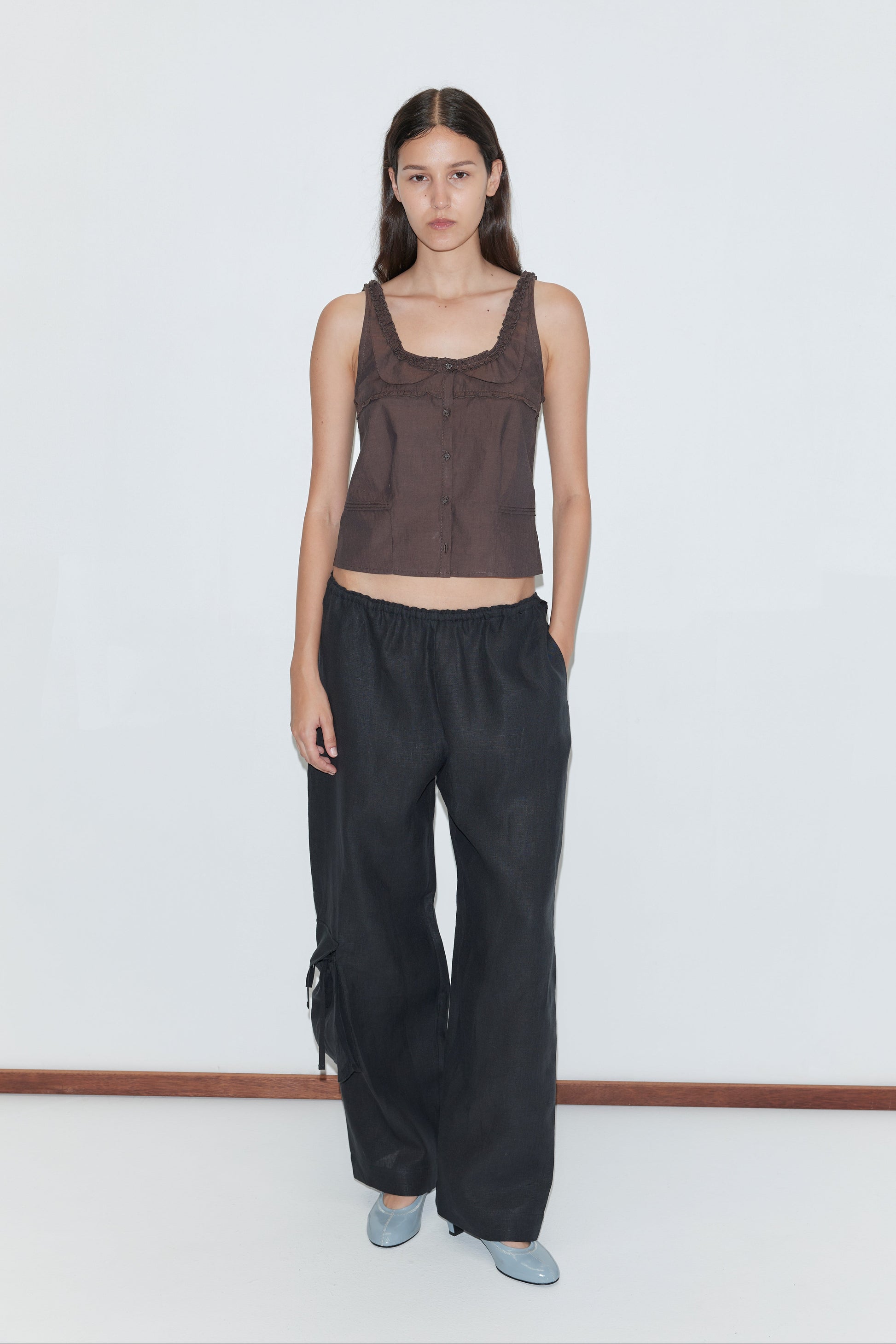 A woman with long dark hair stands against a plain white wall, wearing the Frills Top - Toast by Deiji Studios, loose black pants, and light blue flats. One hand is in her pocket and she has a neutral expression.