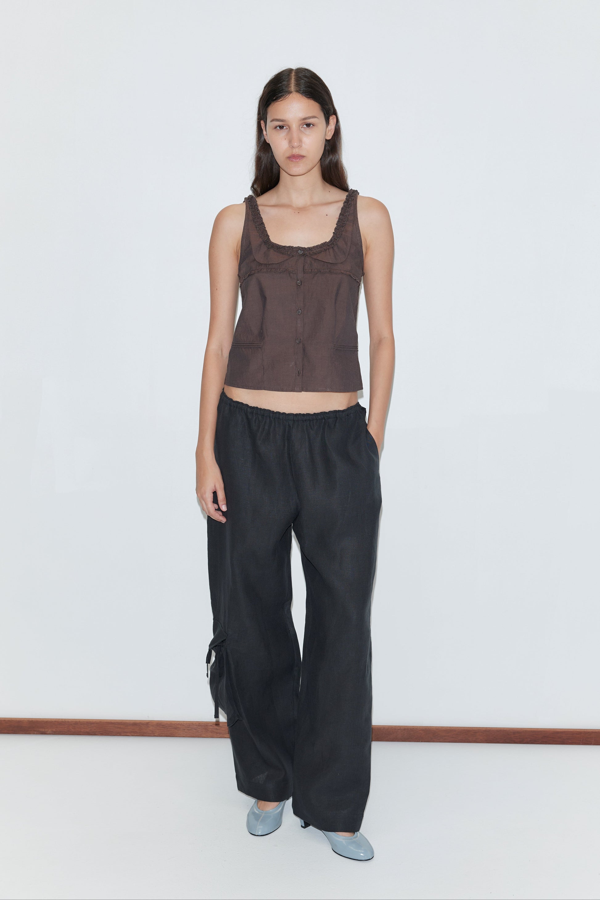 A woman with long dark hair and a neutral expression stands against a white background, wearing Deiji Studios’ Side Pant - Black with an elasticated waistband and her hand in one pocket, styled simply for an effortless look.