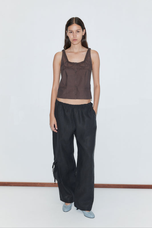 A woman with long dark hair and a neutral expression stands against a white background, wearing Deiji Studios’ Side Pant - Black with an elasticated waistband and her hand in one pocket, styled simply for an effortless look.
