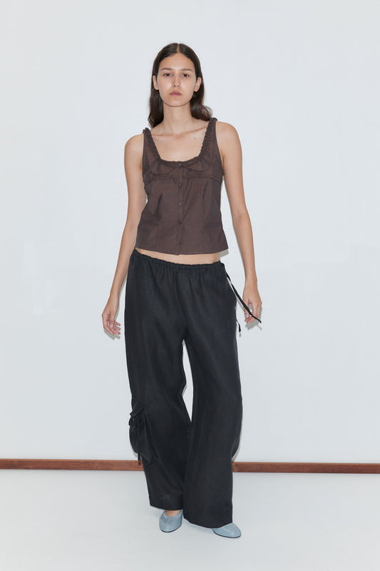 A woman with long brown hair wears a sleeveless brown buttoned top, the Side Pant - Black from Deiji Studios featuring an elasticated waistband, and light blue flats, standing against a plain white background.