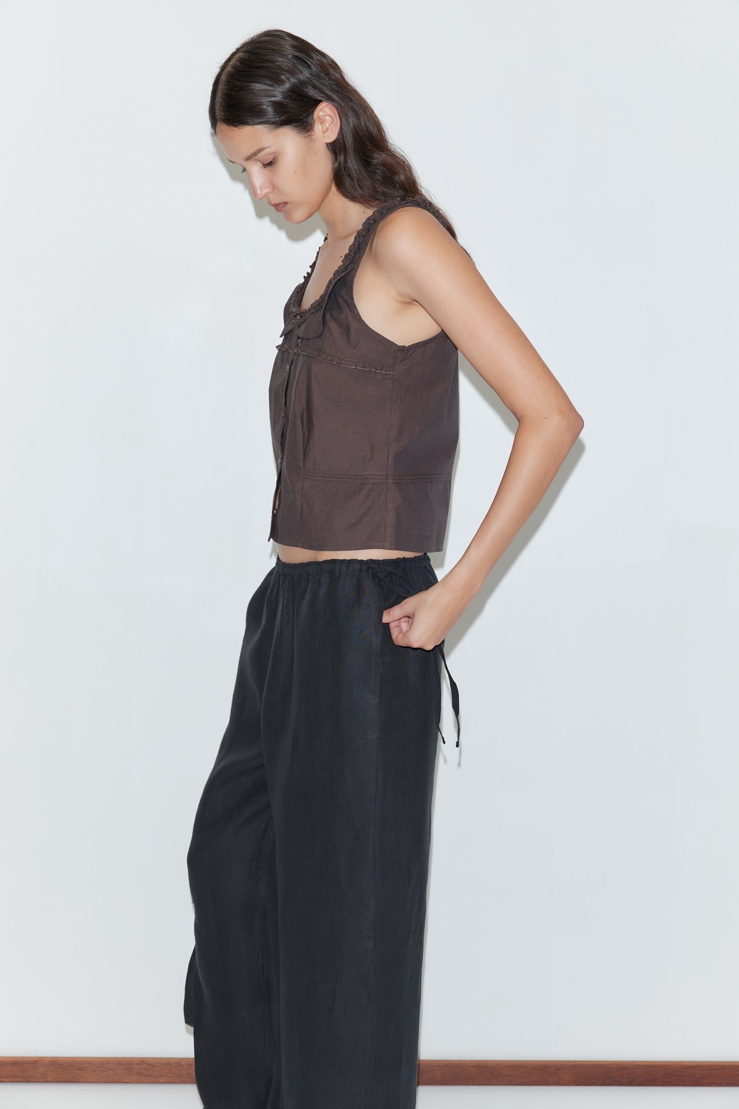A woman with long dark hair stands in profile against a white background, wearing the Frills Top - Toast and loose-fitting black pants, one hand in her pocket.
