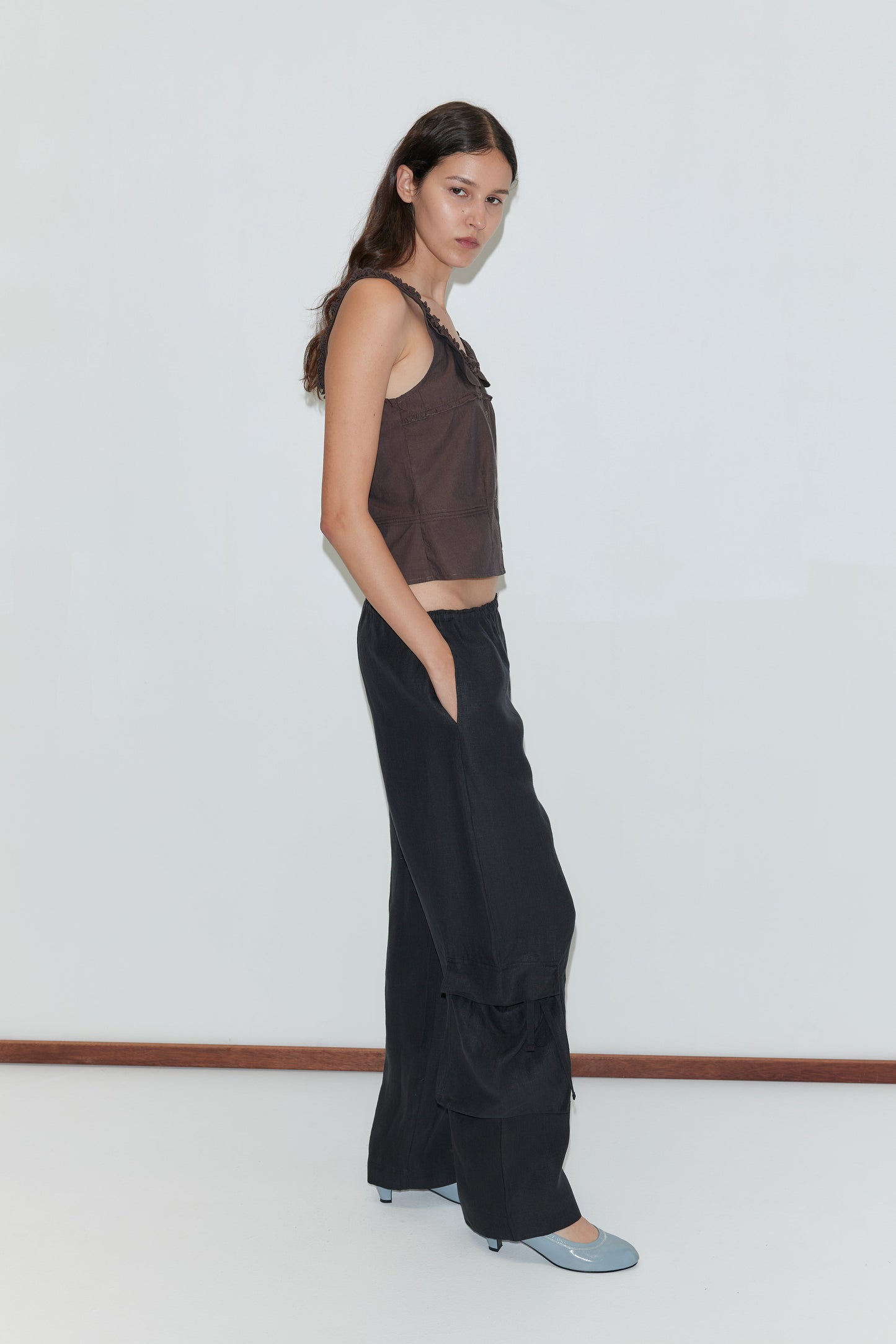 A woman with long brown hair stands against a white wall, wearing a sleeveless brown top and the Side Pant - Black from Deiji Studios. She pairs the elasticated black pants with light blue shoes and looks at the camera with a neutral expression.