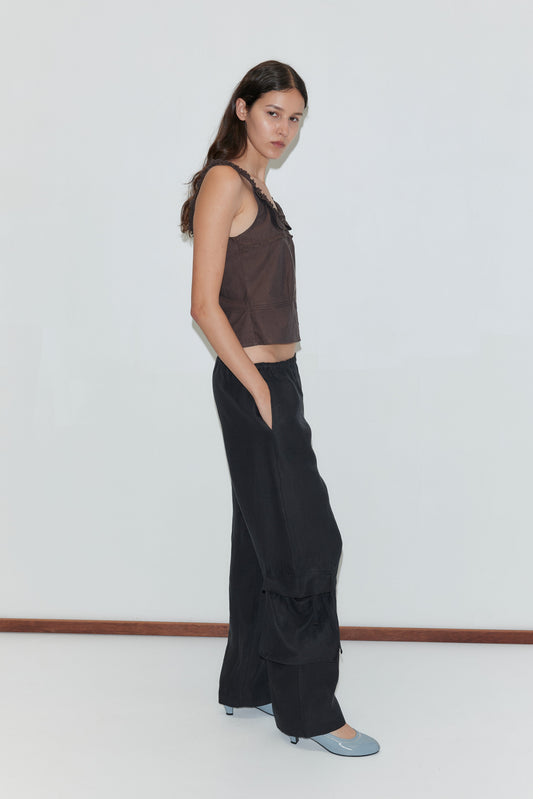 A woman with long brown hair stands against a white wall, wearing a sleeveless brown top and the Side Pant - Black from Deiji Studios. She pairs the elasticated black pants with light blue shoes and looks at the camera with a neutral expression.