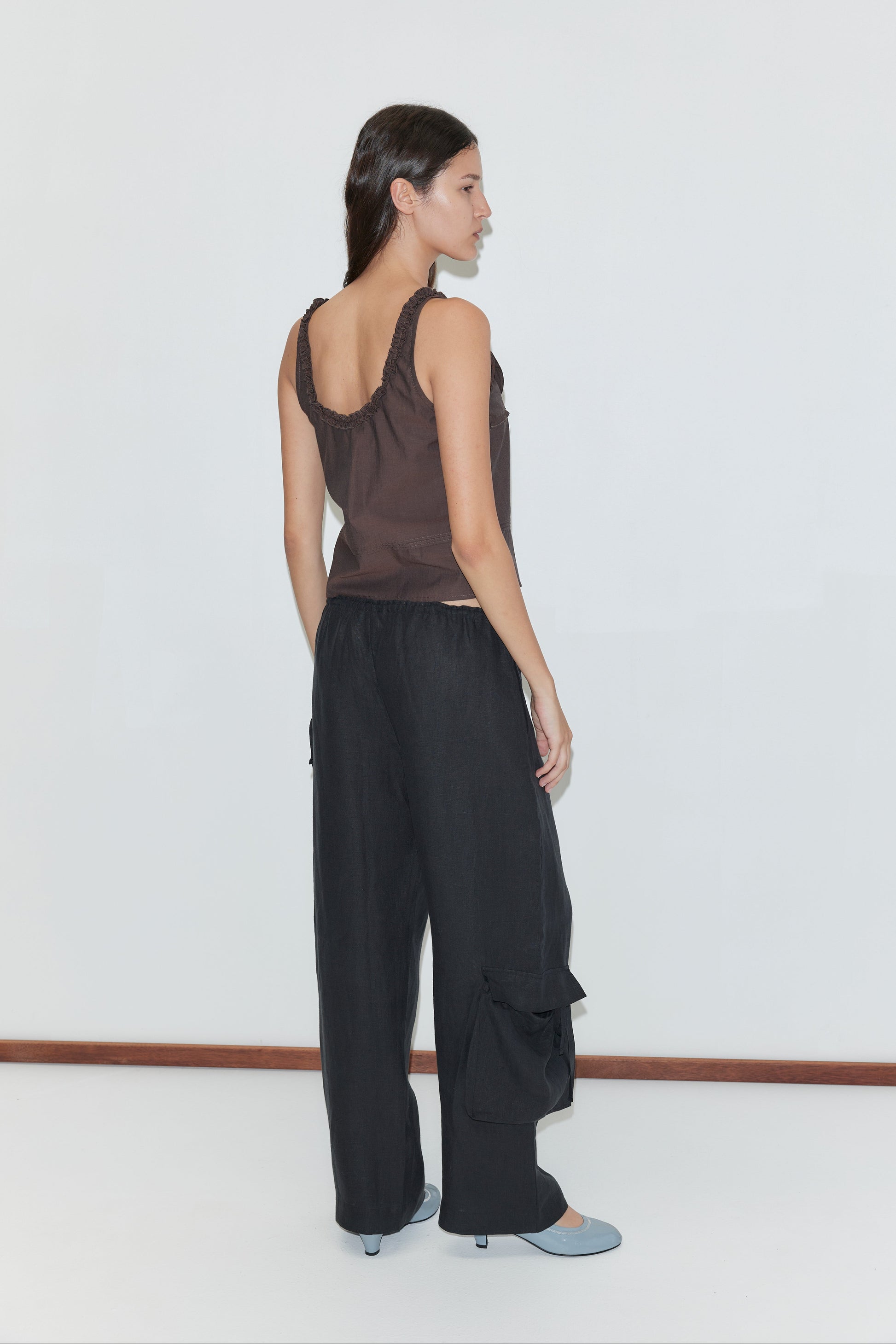 A woman stands sideways against a plain white background, wearing a brown sleeveless top with ruffled straps, the Side Pant in black featuring an elasticated waistband and side pockets, and light blue heeled shoes.