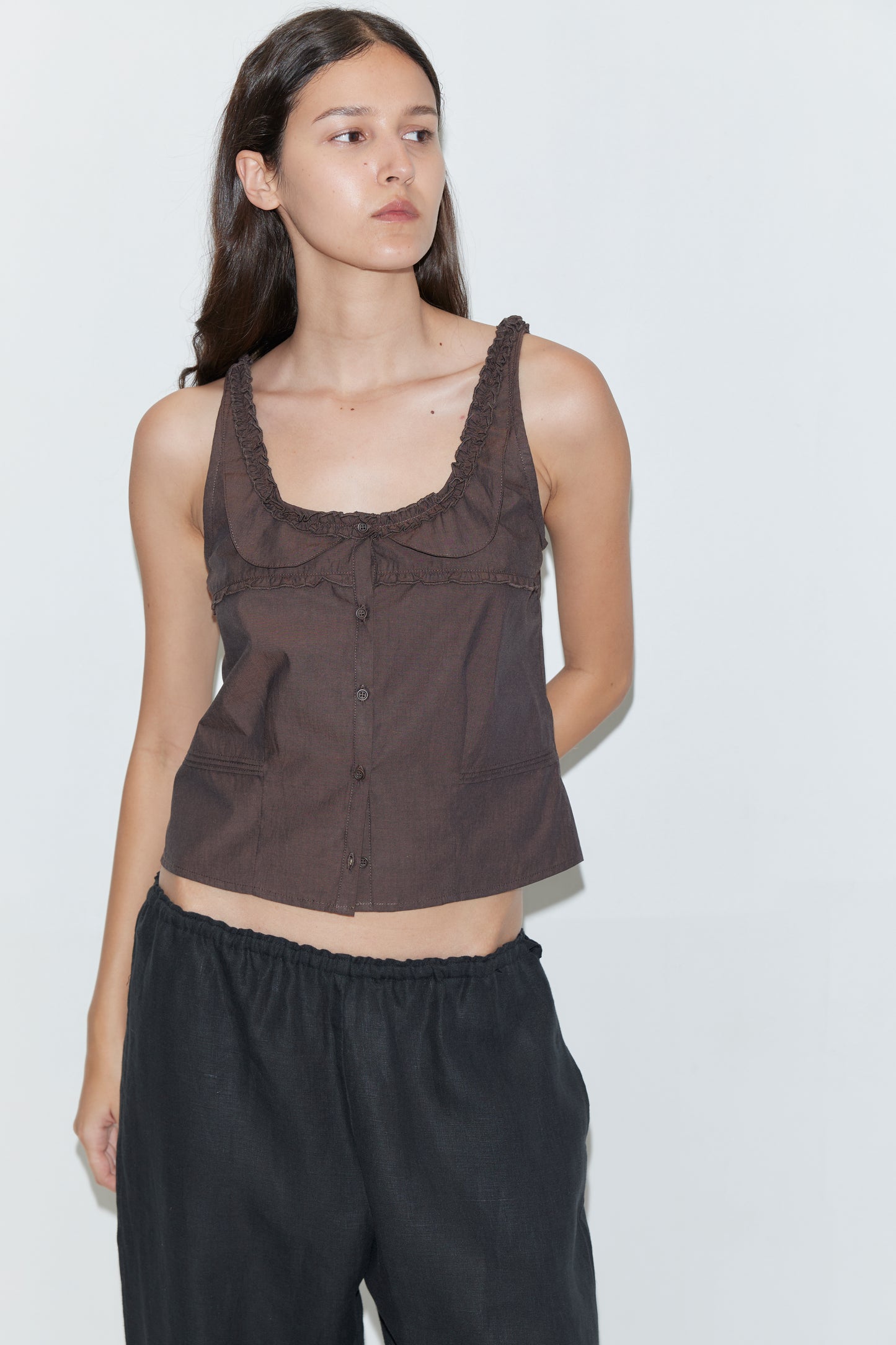 A woman with long dark hair wears the Frills Top - Toast by Deiji Studios and loose black pants, standing against a plain white background and looking to the side with a neutral expression.