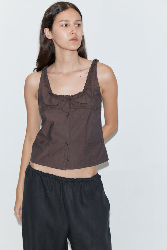 A woman with long dark hair wears the Frills Top - Toast by Deiji Studios and loose black pants, standing against a plain white background and looking to the side with a neutral expression.