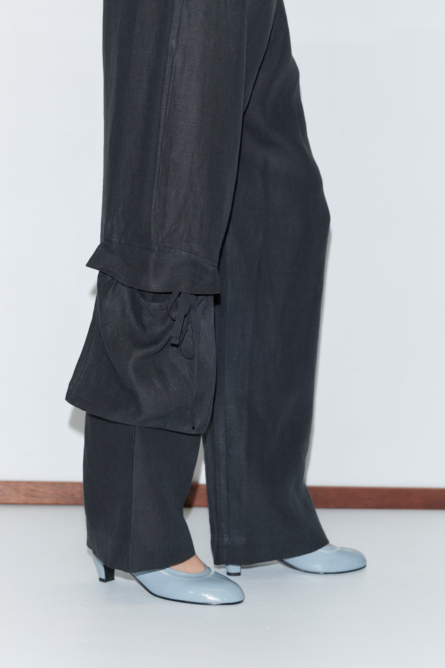 A person wears the Side Pant - Black, featuring wide-leg linen, an elasticated waistband, and tied cuffs, paired with light blue heeled shoes. They stand on a white floor against a white wall with wooden trim.