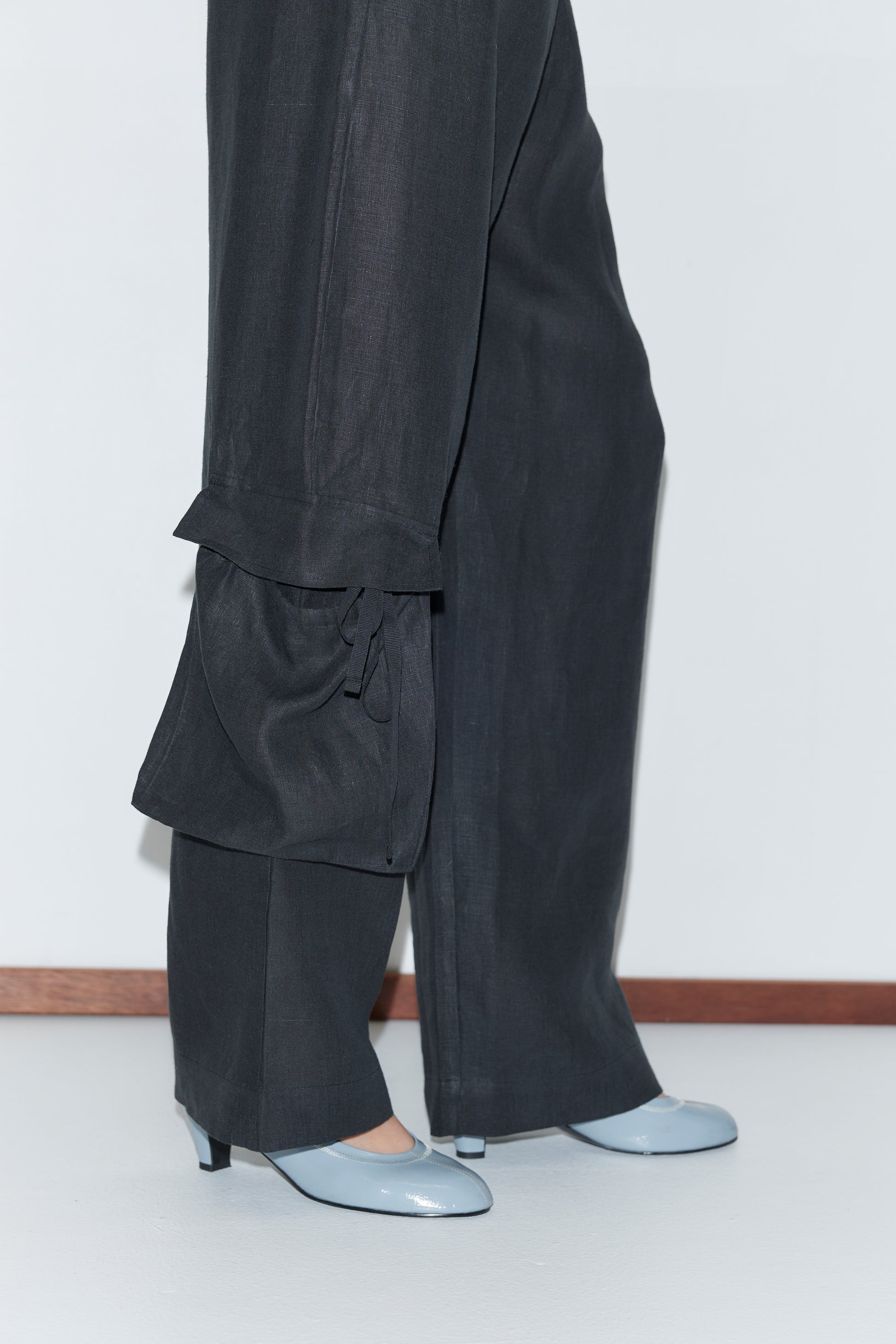 A person wears the Side Pant - Black, featuring wide-leg linen, an elasticated waistband, and tied cuffs, paired with light blue heeled shoes. They stand on a white floor against a white wall with wooden trim.