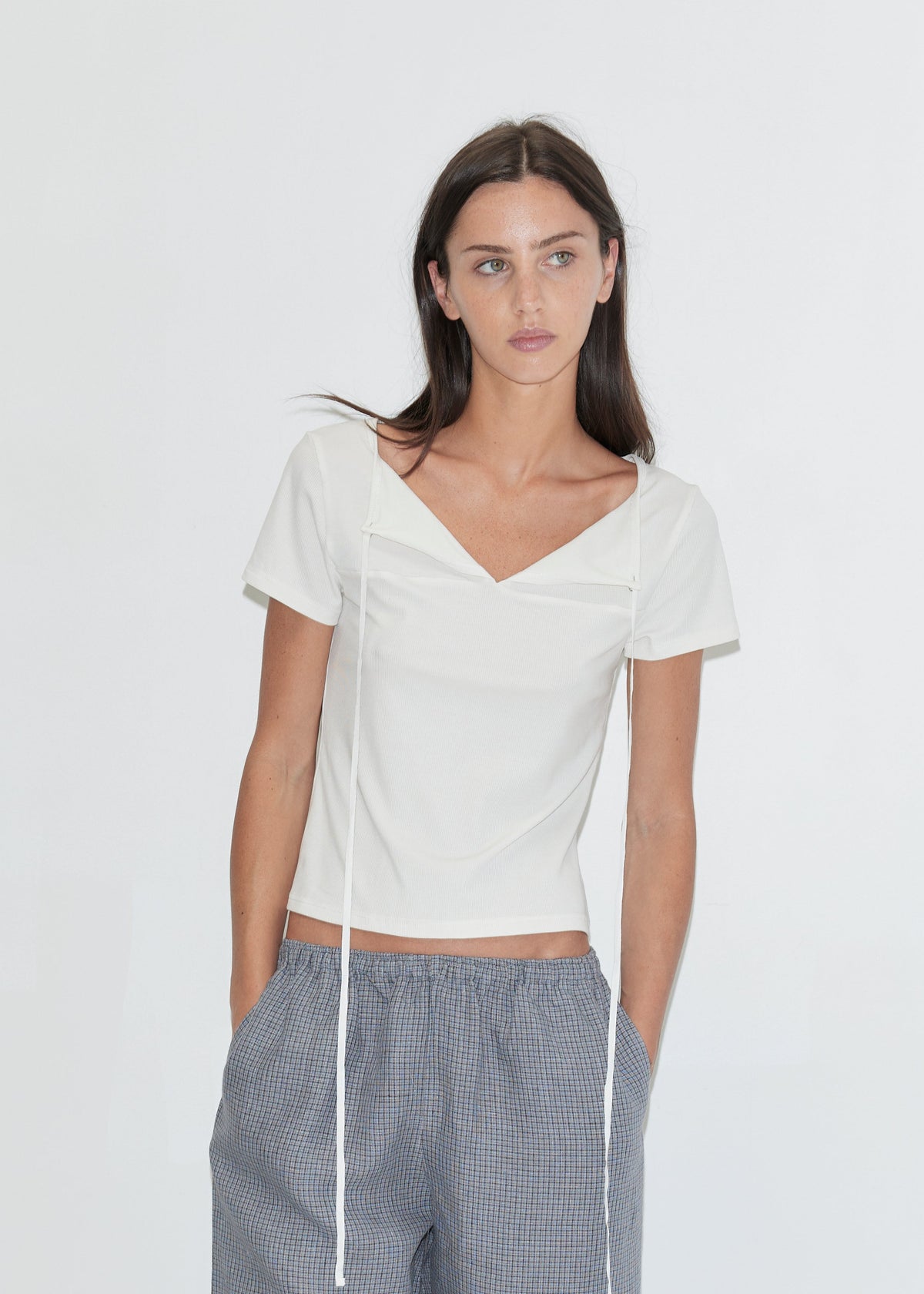 A cropped view of a woman wearing the Linen Ease Trouser - Indigo Check by Deiji Studios, indigo micro-check wide-leg linen trousers with a drawcord waist, and a white tie-back tee against a plain white background.