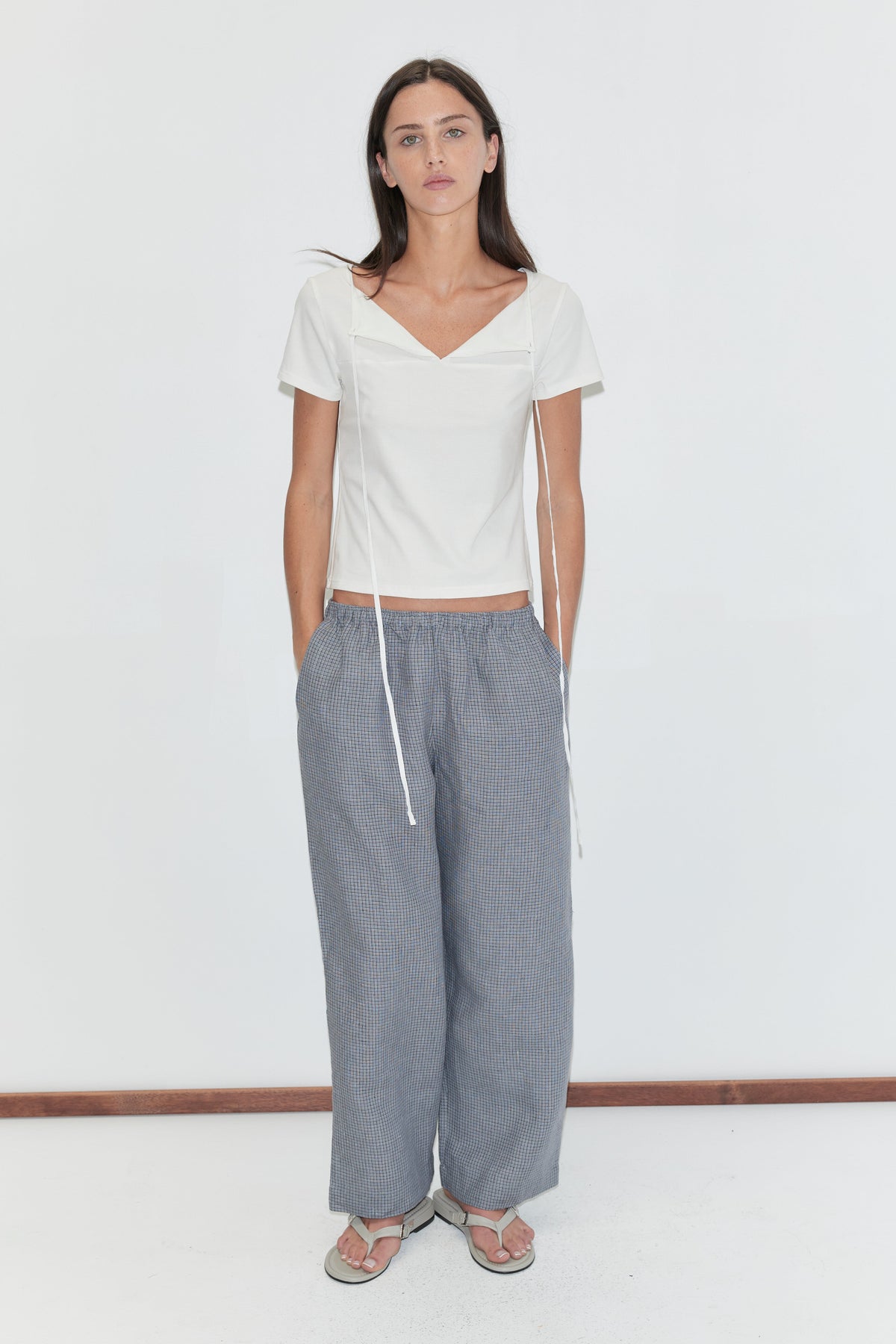 A woman with long dark hair stands against a plain white background with her hands in her pockets, wearing the Linen Ease Trouser - Indigo Check by Deiji Studios, indigo micro-check wide-leg linen trousers, with a white tie-back tee and grey flip-flops.