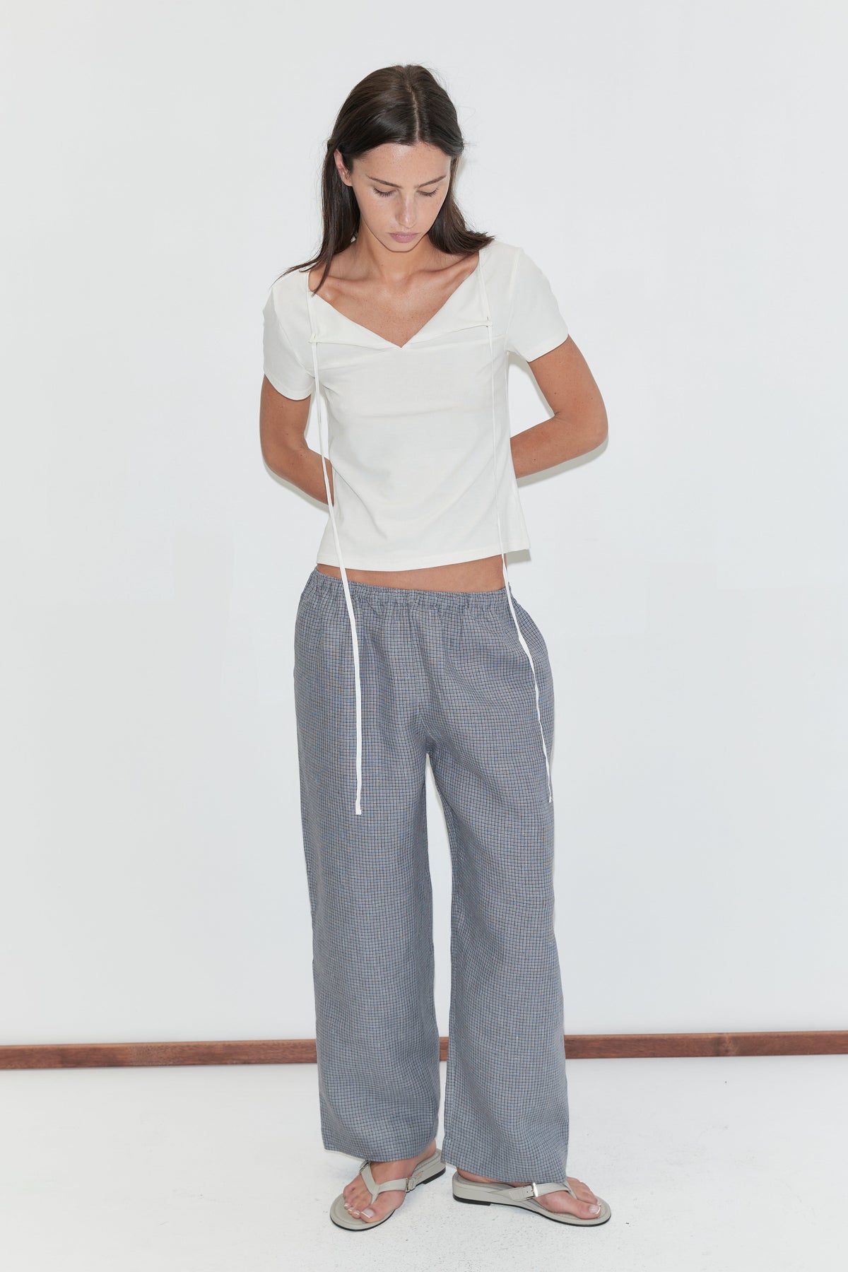 A woman with long dark hair stands against a plain white background looking down, wearing the Linen Ease Trouser - Indigo Check by Deiji Studios, indigo micro-check wide-leg linen trousers, with a white tie-back tee and flip-flops.