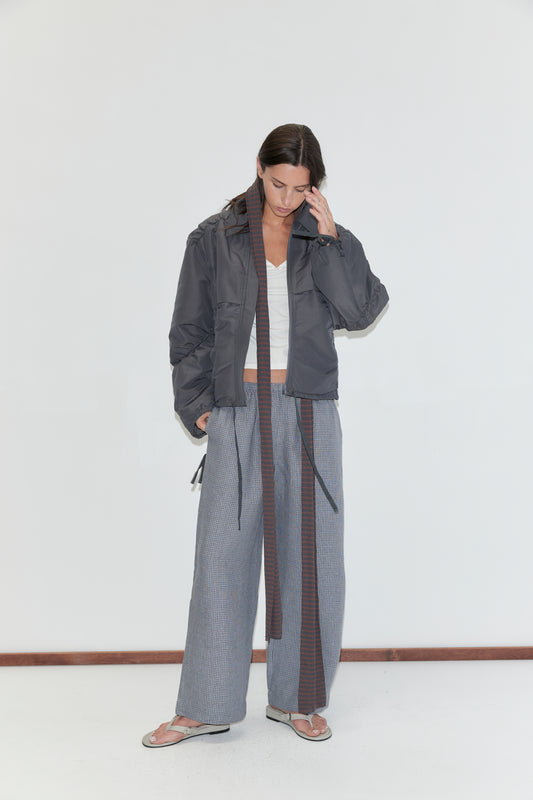 A woman with long dark hair stands against a plain white background wearing the Linen Ease Trouser - Indigo Check by Deiji Studios, indigo micro-check wide-leg linen trousers, with a white top and a slate grey gathered jacket.