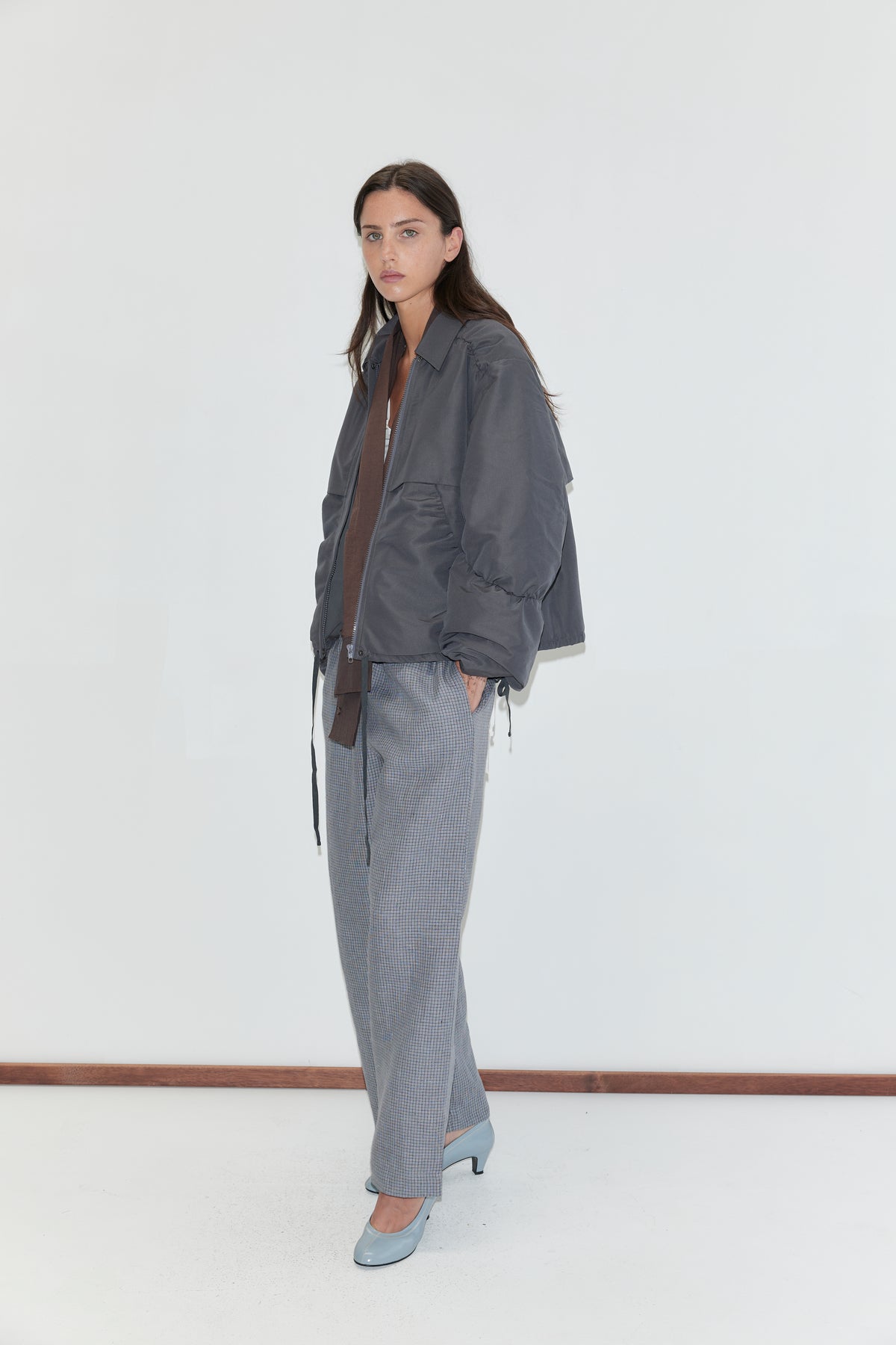 A woman with long dark hair stands against a plain white background wearing the Linen Ease Trouser - Indigo Check by Deiji Studios, indigo micro-check wide-leg linen trousers, with a slate grey gathered jacket and pale blue heels.
