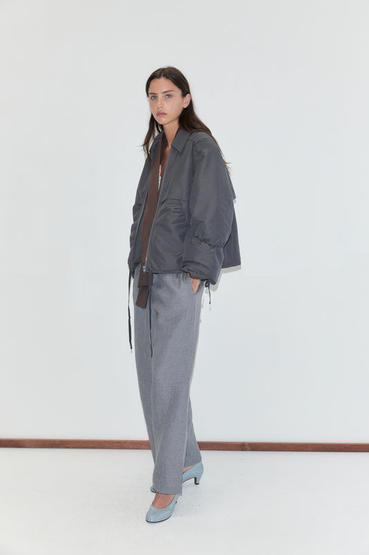 A woman with long dark hair stands against a plain white background wearing the Linen Ease Trouser - Indigo Check by Deiji Studios, indigo micro-check wide-leg linen trousers, with a slate grey gathered jacket and pale blue heels.