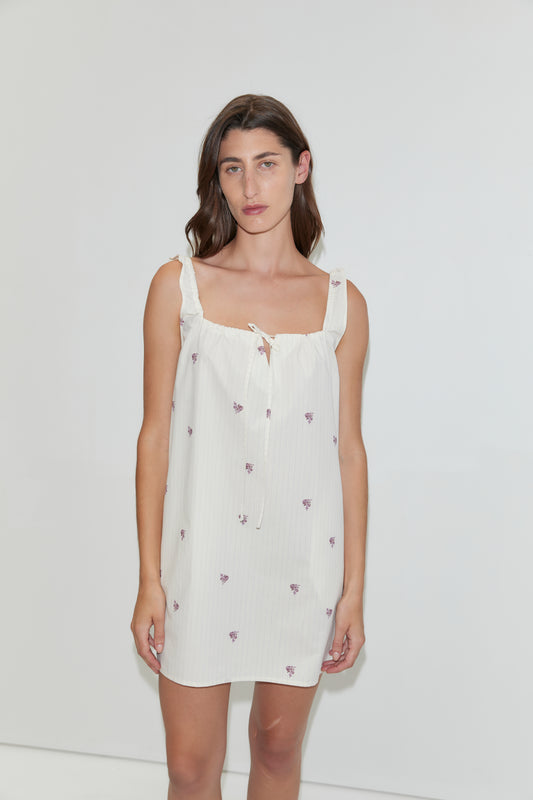 A woman stands against a plain white background wearing the Bow Dress - Floral Stripe, a white mini-length dress featuring small purple floral prints and ruffled shoulder straps.