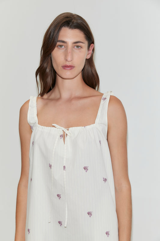 A woman with long brown hair wears the Bow Dress - Floral Stripe, a white mini-length dress featuring small purple flower prints, a bow at the neckline, and stands against a plain light background.
