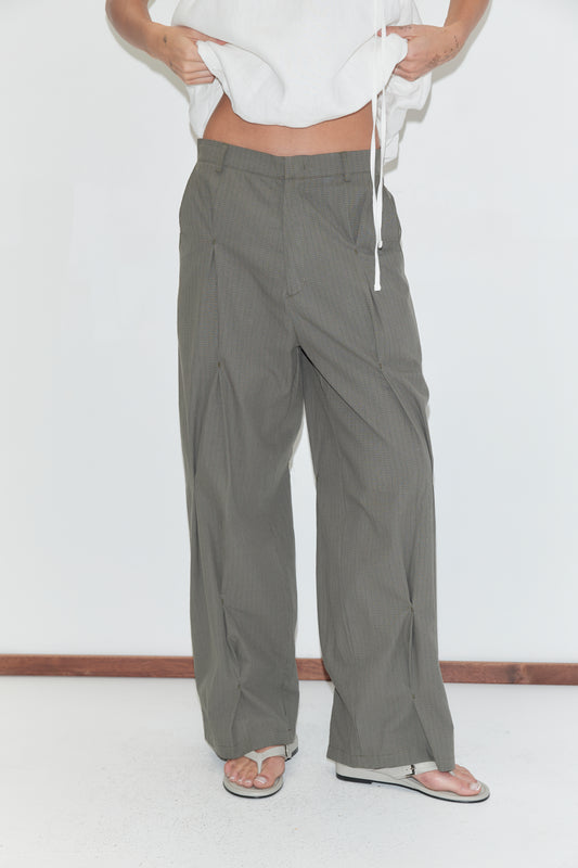 A cropped view of the Multi Pleat Pant - Moss Stripe by Deiji Studios, moss-green fine-check wide-leg pleated trousers with a waistband, worn with a white top and flip-flops against a plain white background.