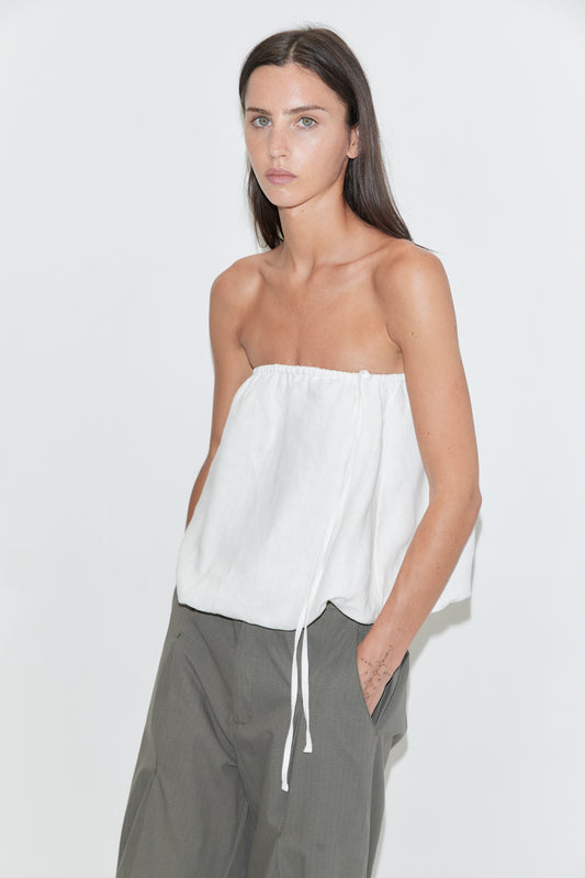 A woman with long dark hair stands against a plain white background wearing the Bubble Top - White by Deiji Studios, a strapless white linen top with a gathered edge and drawcord side tie, paired with olive check wide-leg trousers.