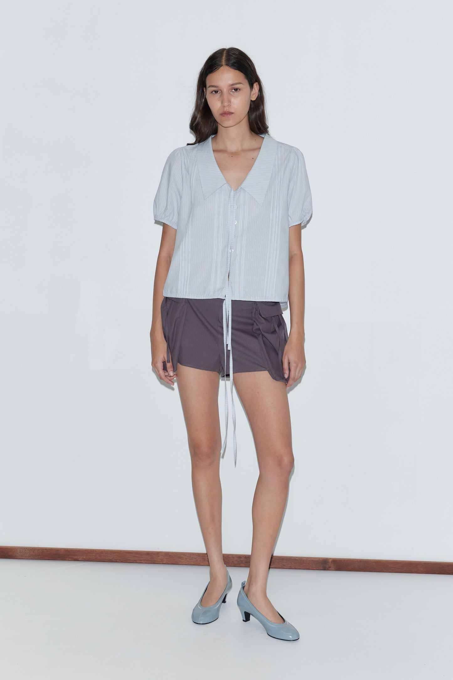 A woman with long brown hair and a neutral expression stands against a white wall, wearing the Fine Collar Top - Mist Stripe, dark gray shorts, and light blue low-heeled shoes.