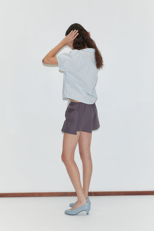 A woman stands sideways in front of a white wall, wearing a Deiji Studios light blue shirt, Micro Pocket Short - Mulberry Stripe, and blue heels. She touches her hair with both hands while looking slightly downward.
