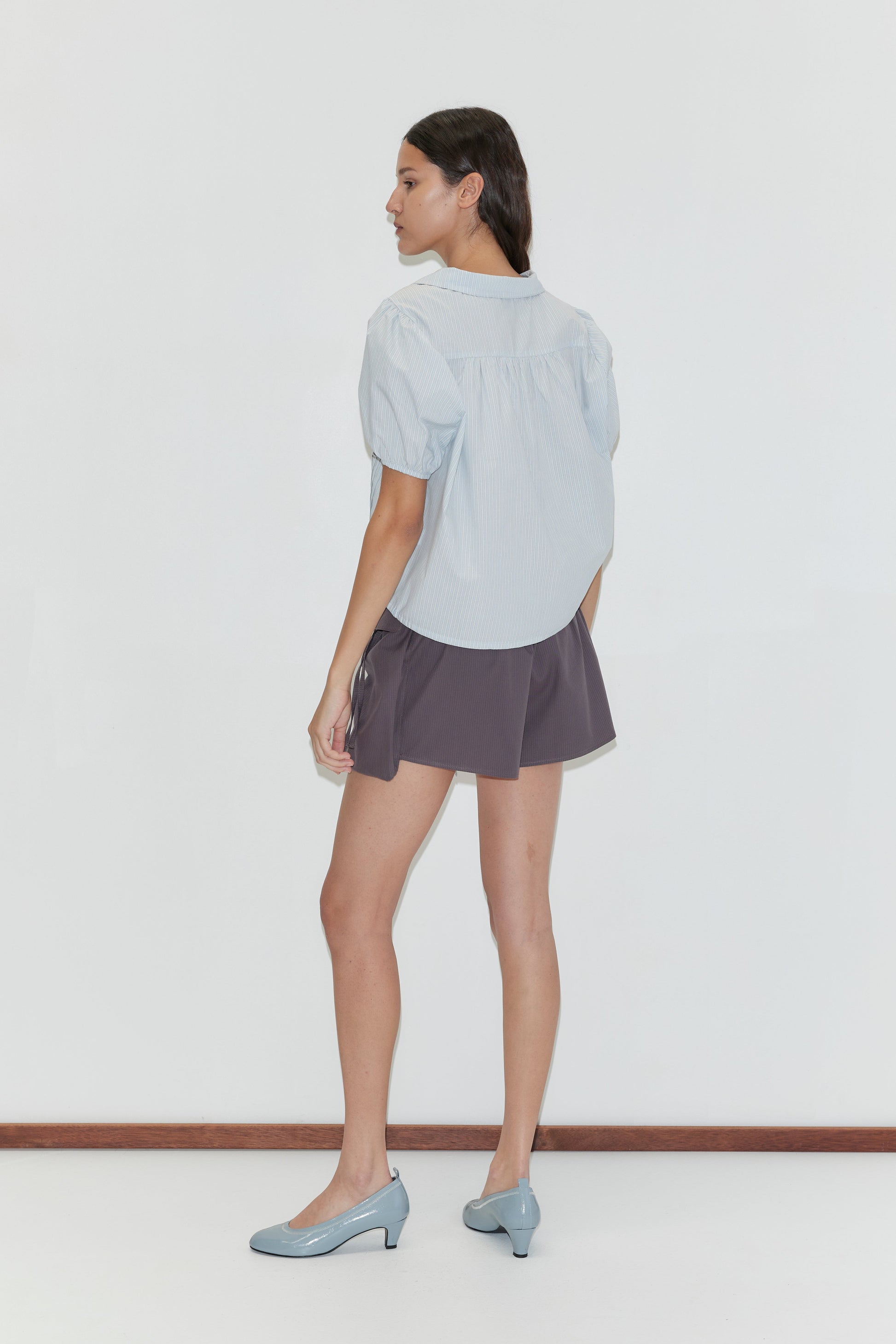 A woman with long hair and a neutral expression stands sideways against a white background, wearing the Fine Collar Top - Mist Stripe, gray shorts, and light blue low-heeled shoes.