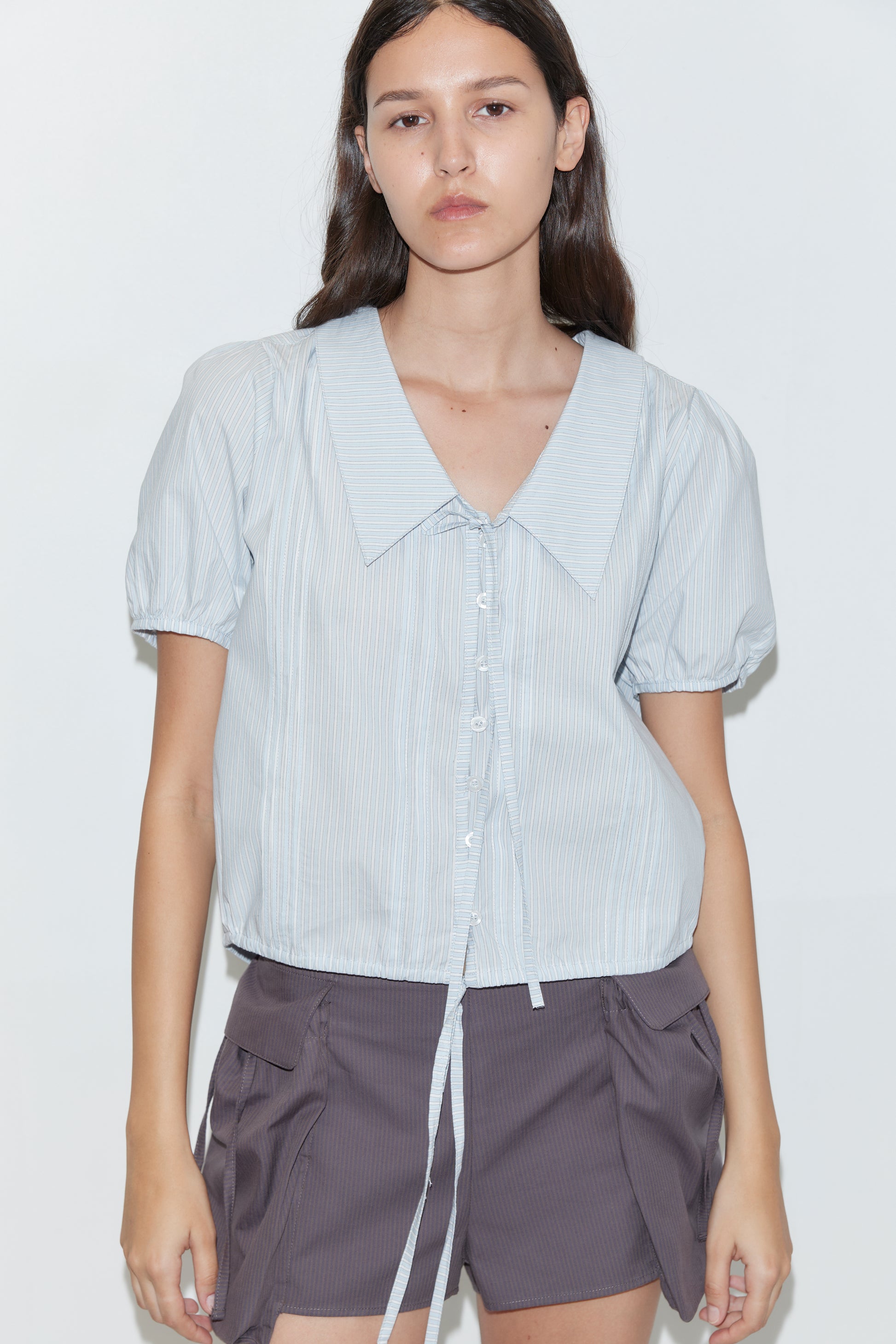 A person with long brown hair wears the deiji studios Fine Collar Top in Mist Stripe (light blue cotton poplin, short sleeves, tie detail), paired with dark gray large pocket shorts. The background is plain white.