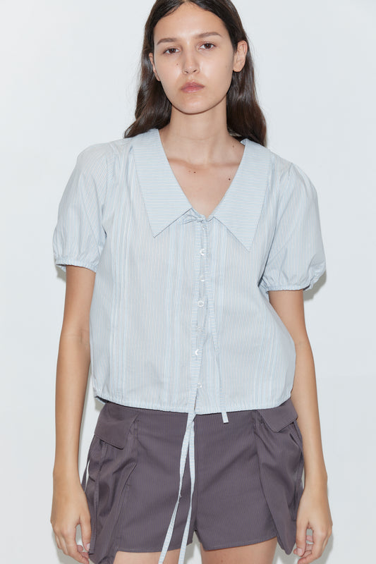 A person with long brown hair wears the deiji studios Fine Collar Top in Mist Stripe (light blue cotton poplin, short sleeves, tie detail), paired with dark gray large pocket shorts. The background is plain white.