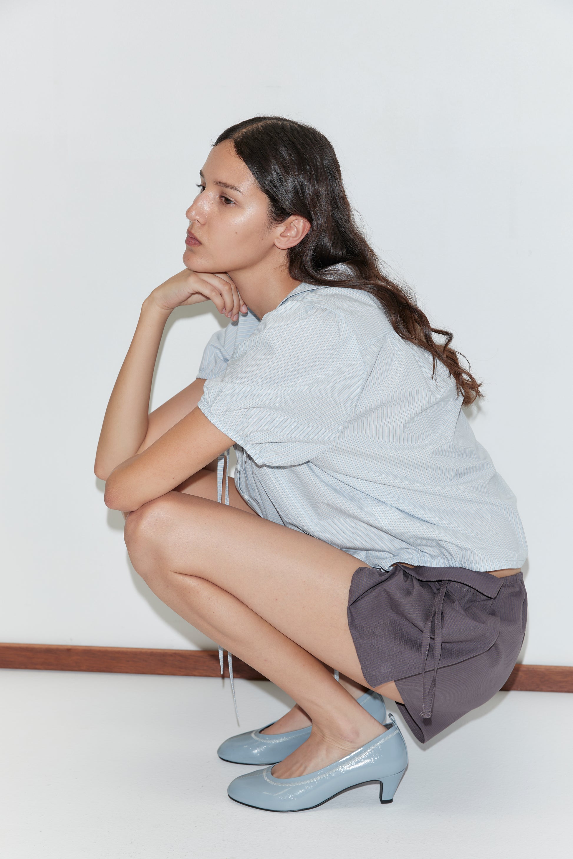 A woman with long brown hair, wearing the Fine Collar Top - Mist Stripe from Deiji Studios, dark gray shorts, and light blue heels, squats with one hand on her chin against a plain white background.
