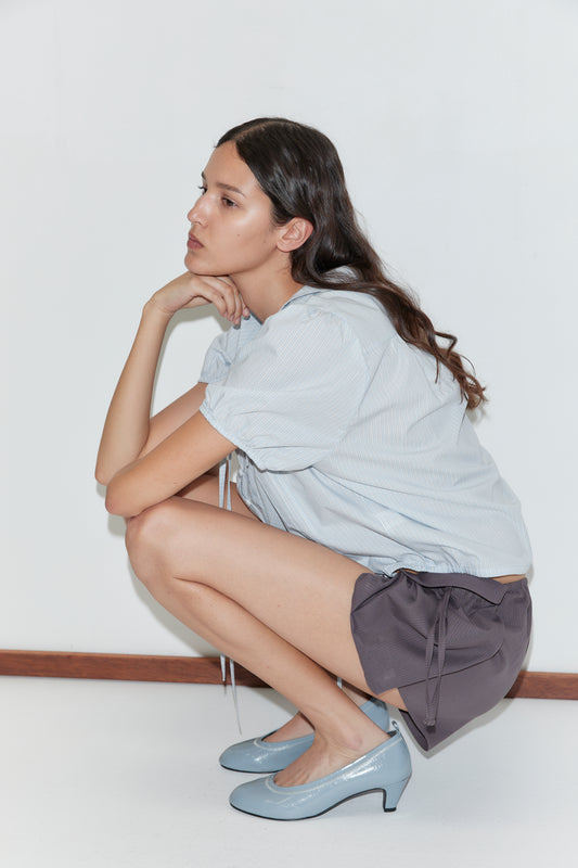 A woman with long brown hair, wearing the Fine Collar Top - Mist Stripe from Deiji Studios, dark gray shorts, and light blue heels, squats with one hand on her chin against a plain white background.