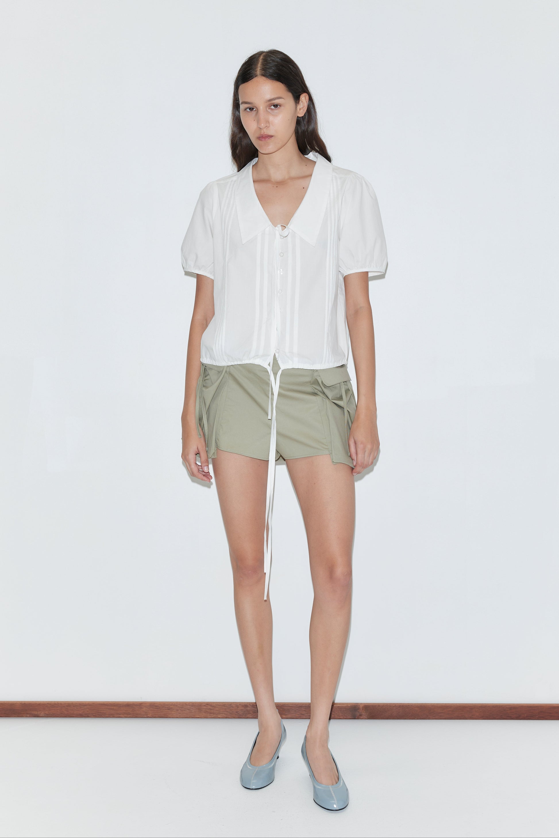 A woman with straight dark hair stands against a white background wearing the Fine Collar Top - White by Deiji Studios with a collared V-neck and drawstring, paired with light olive green shorts and light blue flats.