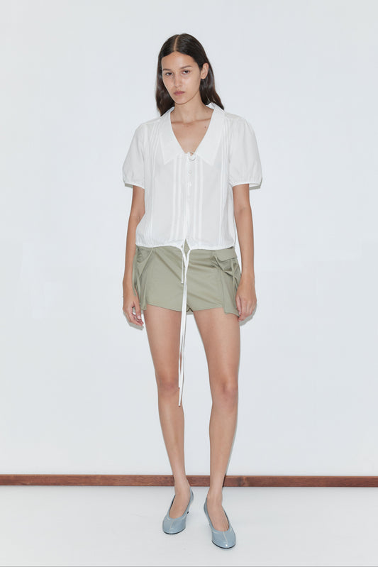 A woman with straight dark hair stands against a white background wearing the Fine Collar Top - White by Deiji Studios with a collared V-neck and drawstring, paired with light olive green shorts and light blue flats.