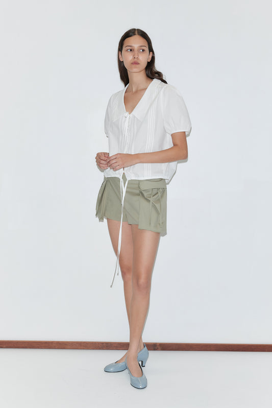 A woman stands against a plain white background wearing the Fine Collar Top - White by Deiji Studios with light green drawstring shorts and light blue low-heeled shoes.