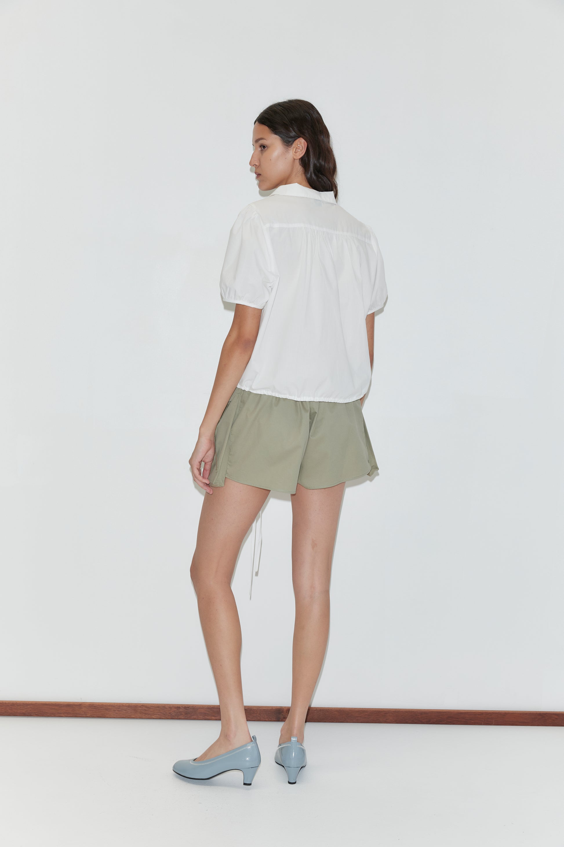 A woman stands against a white background, facing slightly away. She wears the Fine Collar Top - White from Deiji Studios, paired with light green shorts and light blue heels. Her dark hair is pulled back near a wooden floorboard.