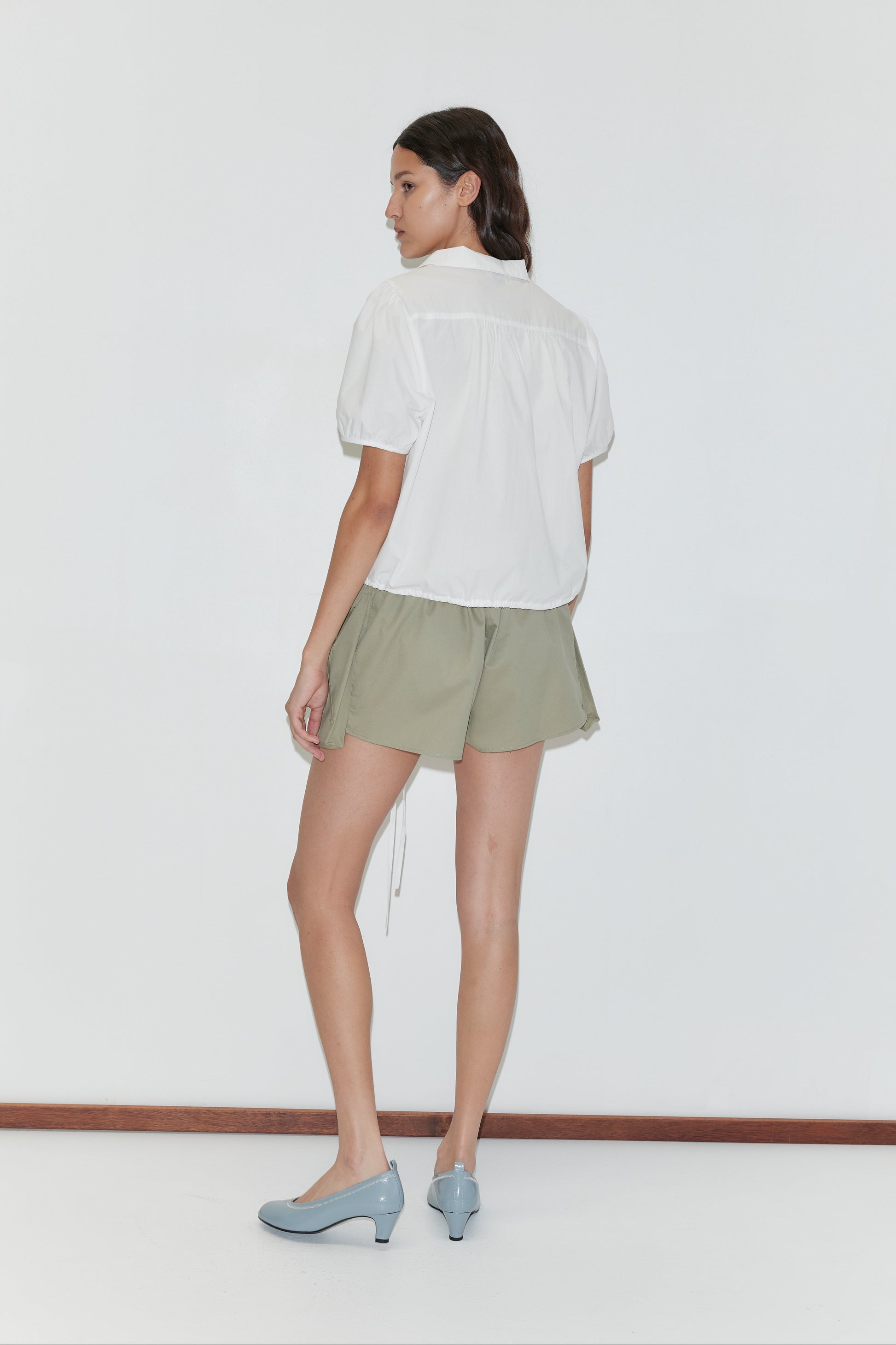 A woman stands against a plain white wall, facing away. She wears a white short-sleeve blouse, the Micro Pocket Short in Stone Green by Deiji Studios, light blue low-heeled shoes, and her hair is styled in a loose braid.