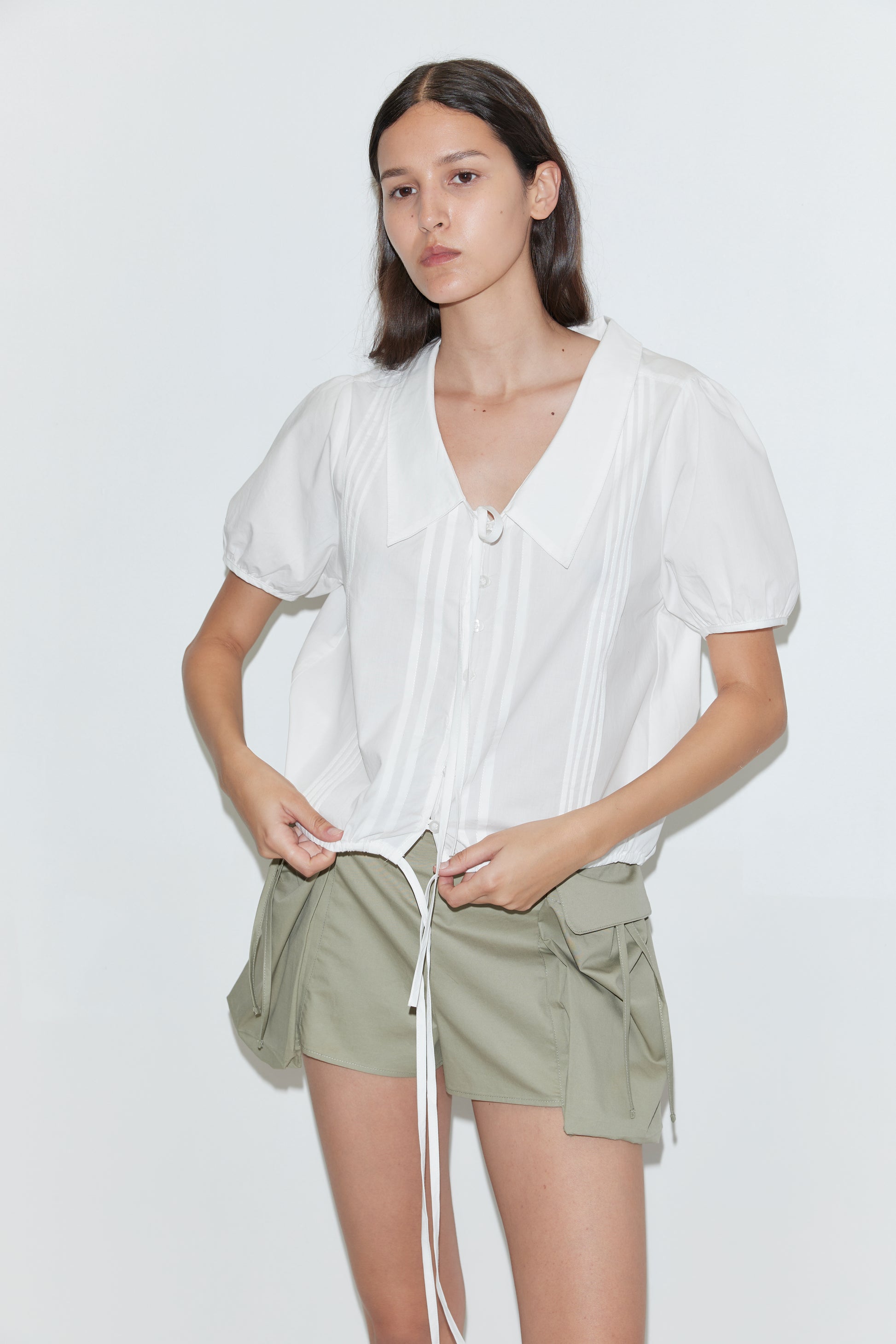 A person wearing the Deiji Studios Fine Collar Top in white cotton poplin with a wide collared v-neck and green shorts with pockets stands against a plain white background.