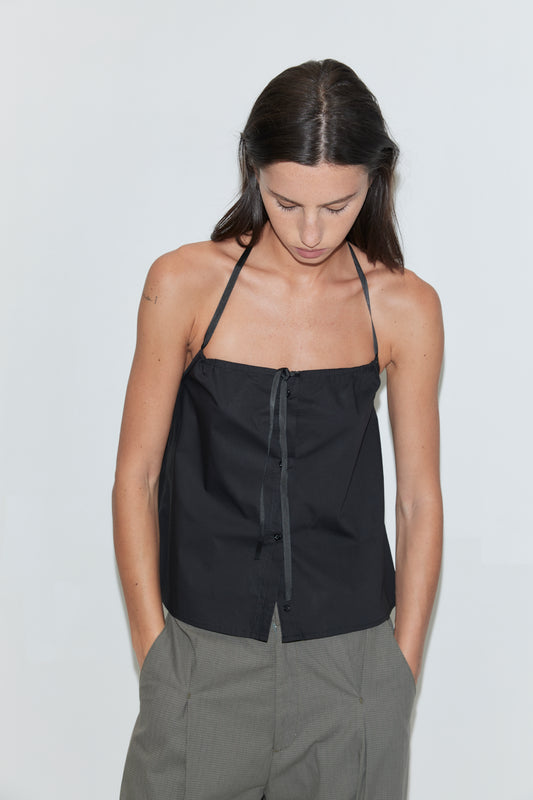 A cropped view of a woman wearing the Simple Tie Top - Black by Deiji Studios, a black halter top with a gathered neckline and drawstring front tie, with grey check trousers against a plain white background.
