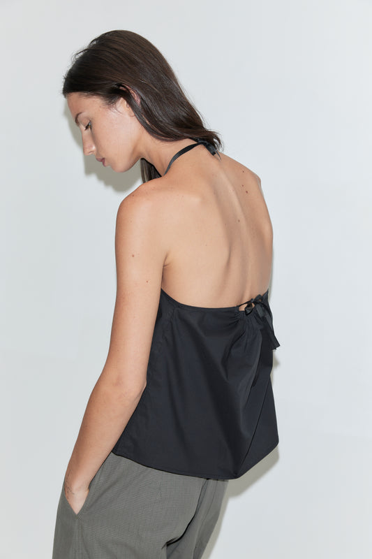 A woman with long dark hair stands in profile against a plain white background wearing the Simple Tie Top - Black by Deiji Studios, a black halter top with an open back and tie fastening, with grey check trousers.