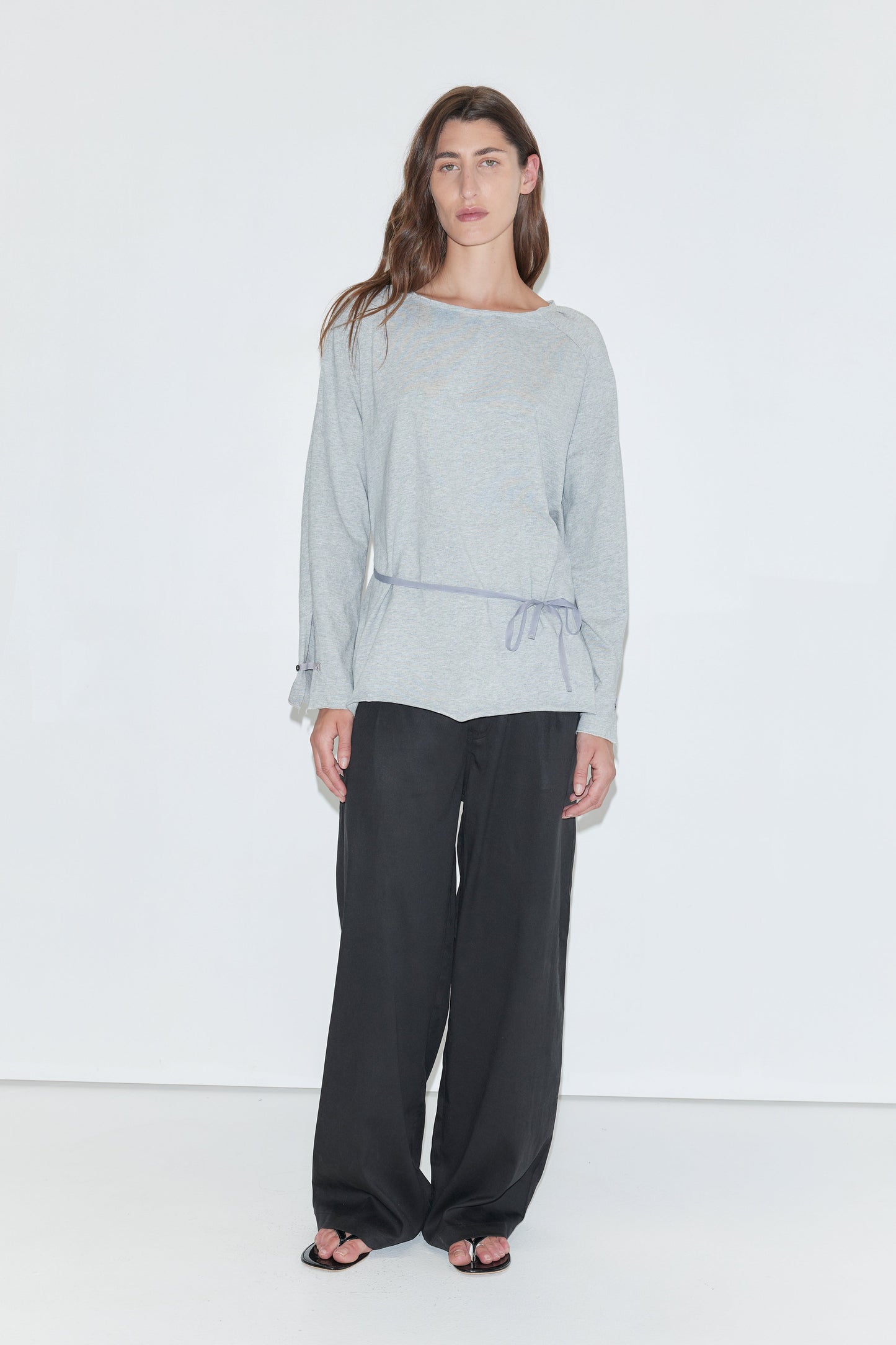 A woman with long dark hair stands against a plain white background wearing the Wided Longsleeve - Grey Marle by Deiji Studios, a grey marle jersey top with a wide neckline and drawstring hem, with black wide-leg trousers and sandals.