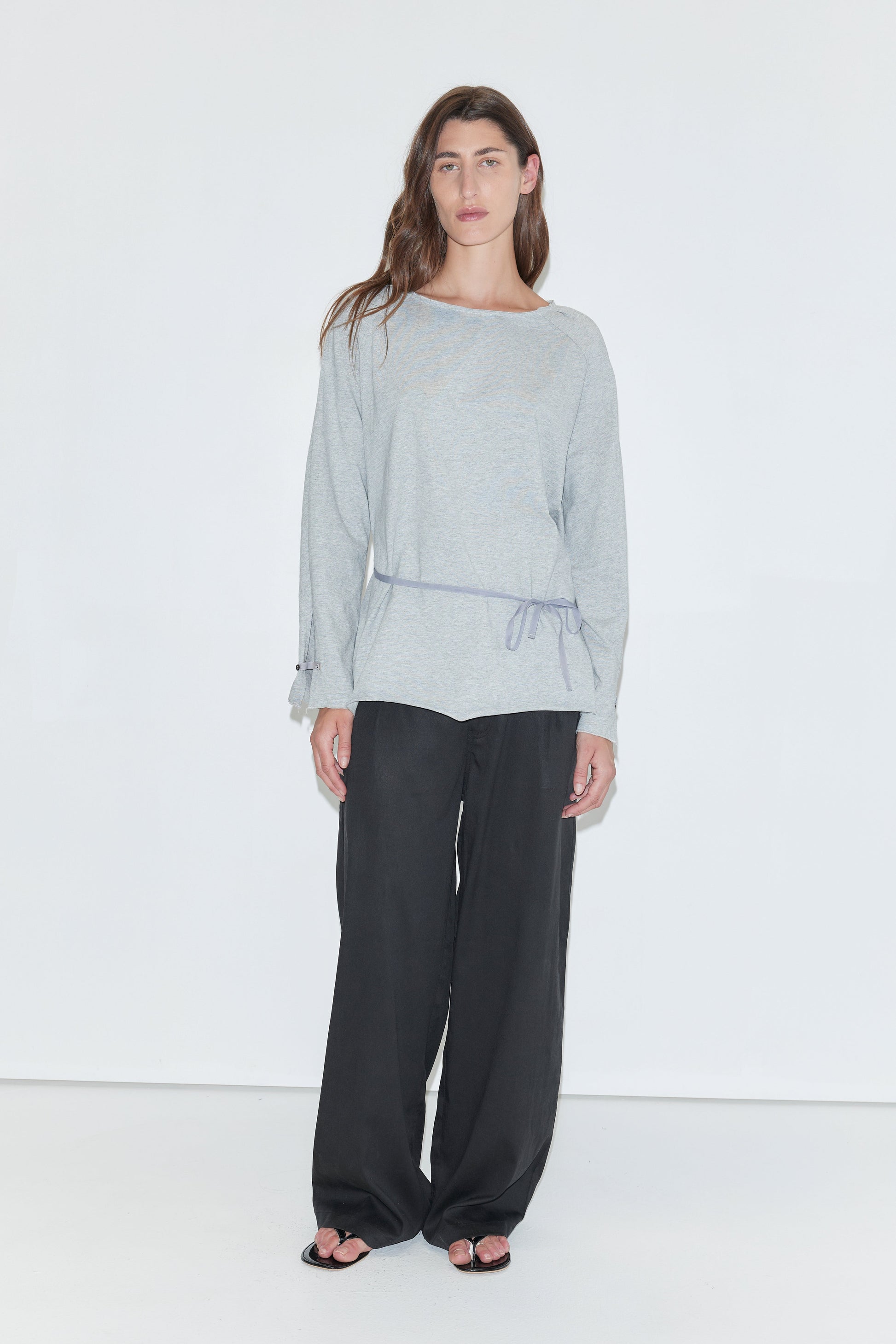 A woman with long dark hair stands against a plain white background wearing the Wided Longsleeve - Grey Marle by Deiji Studios, a grey marle jersey top with a wide neckline and drawstring hem, with black wide-leg trousers and sandals.