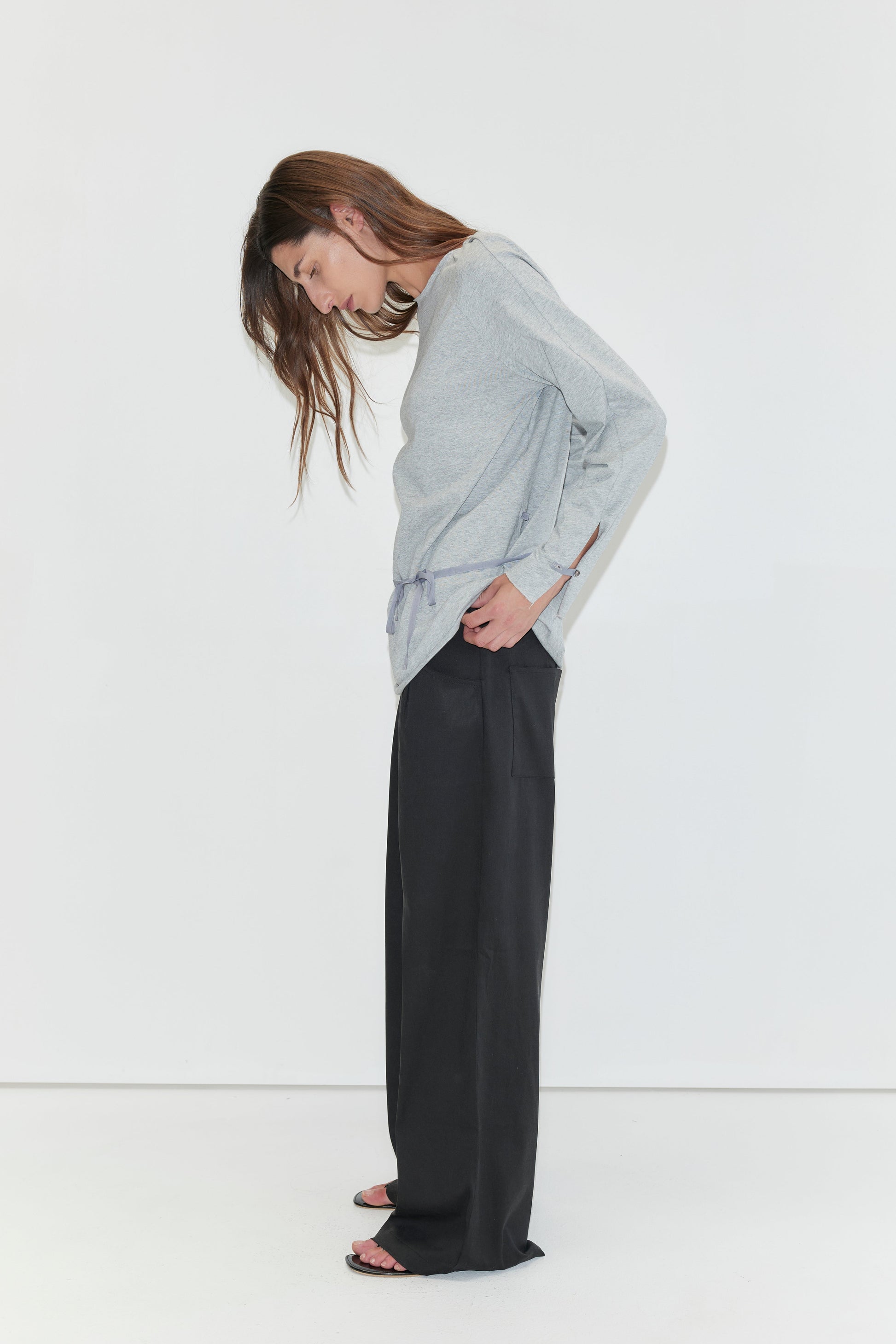 A woman with long dark hair stands in profile against a plain white background wearing the Wided Longsleeve - Grey Marle by Deiji Studios, a grey marle jersey top with a drawstring hem, with black wide-leg trousers.