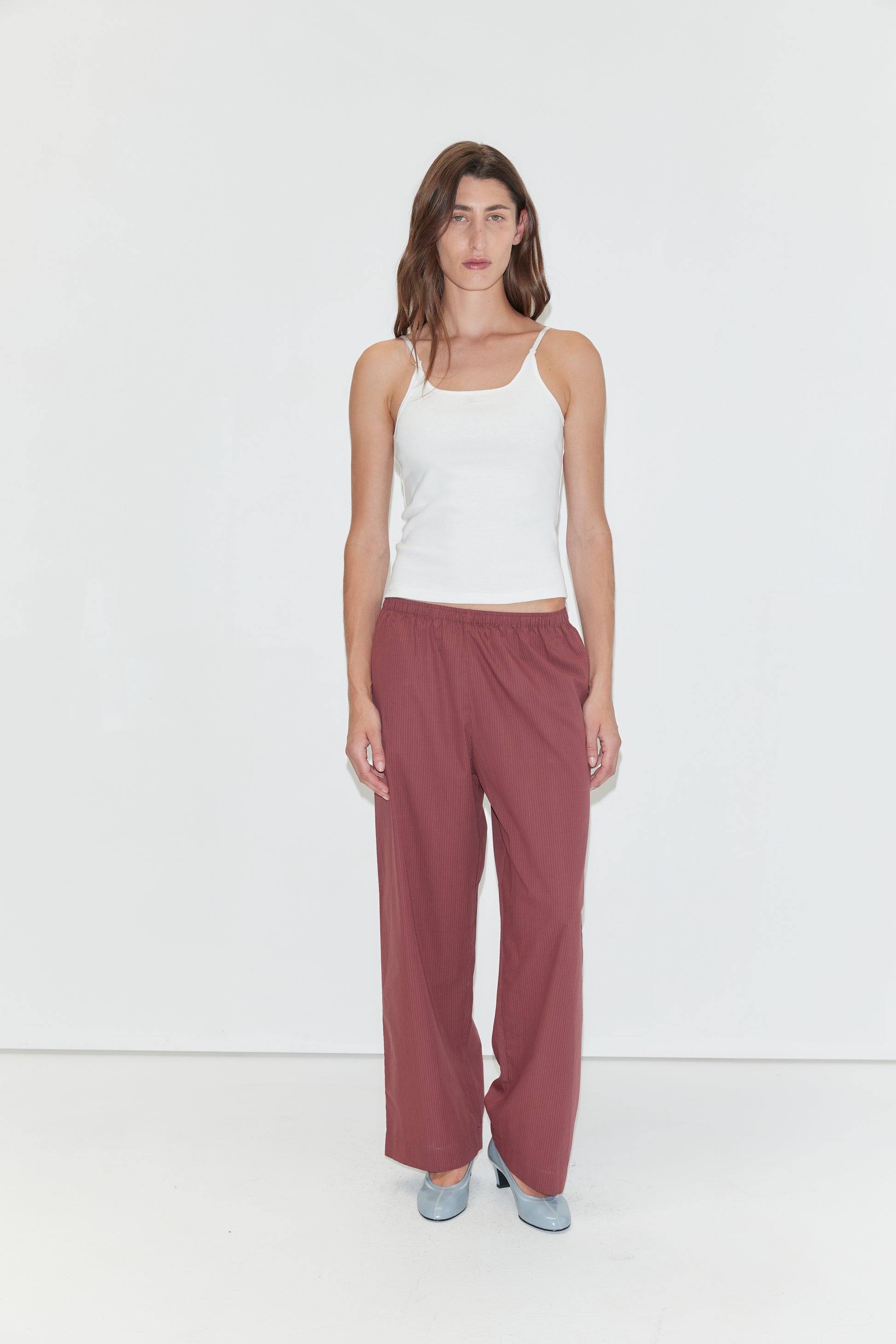 A woman with long brown hair and a neutral expression stands against a plain white background, wearing a white tank top and the Ease Trouser - Rust Stripe by Deiji Studios. She pairs the outfit with light blue shoes.