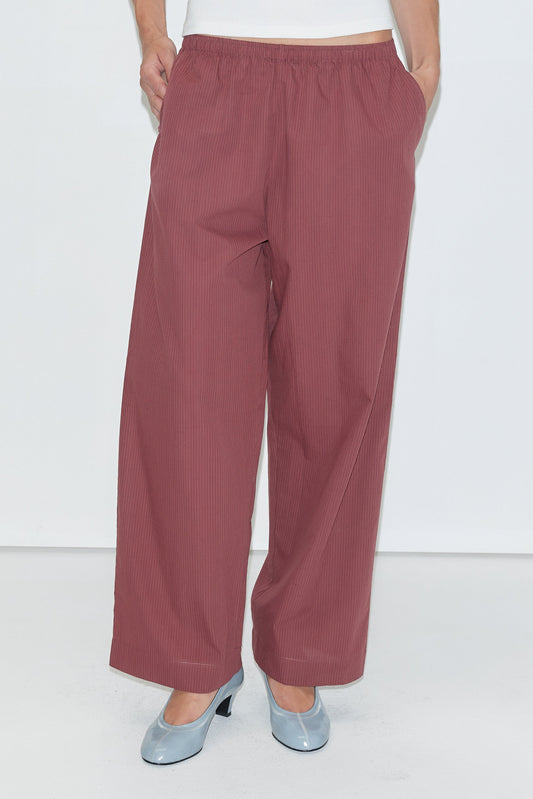 A person wearing the Ease Trouser - Rust Stripe from Deiji Studios with hands in pockets, paired with light blue heeled shoes against a plain white background.